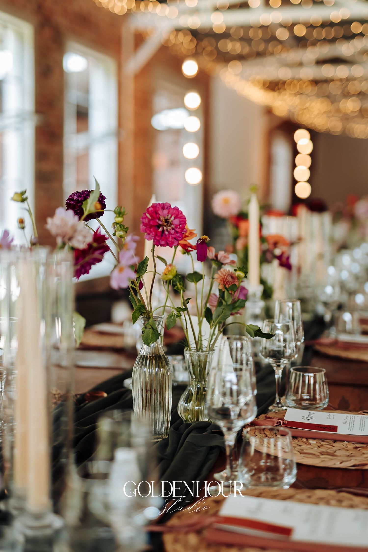 Elegant wedding reception table with pink dahlias in glass vases, crystal glasses, and warm string lights overhead.