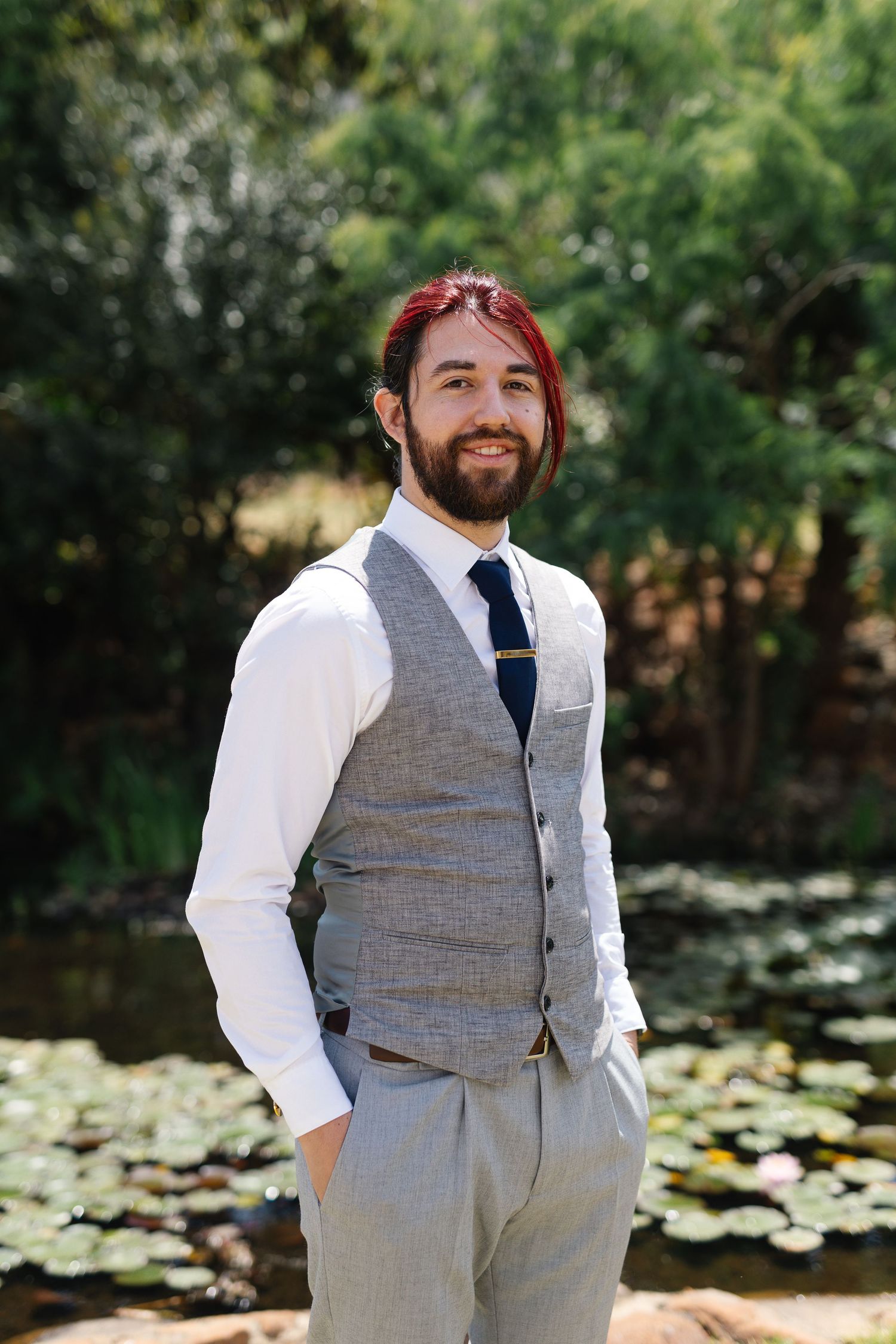 A person wearing a grey vest and navy tie smiles outdoors by a pond with lily pads in the background.