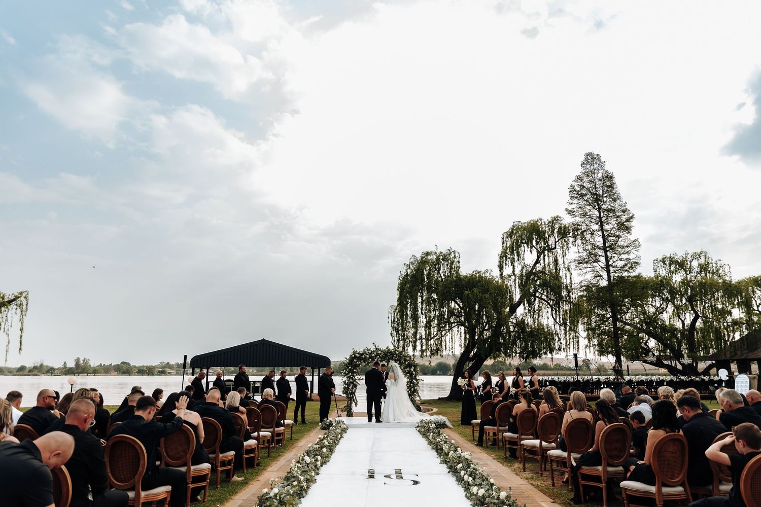 A panoramic view of an outdoor wedding ceremony setup with rustic wooden chairs and a lakeside backdrop.