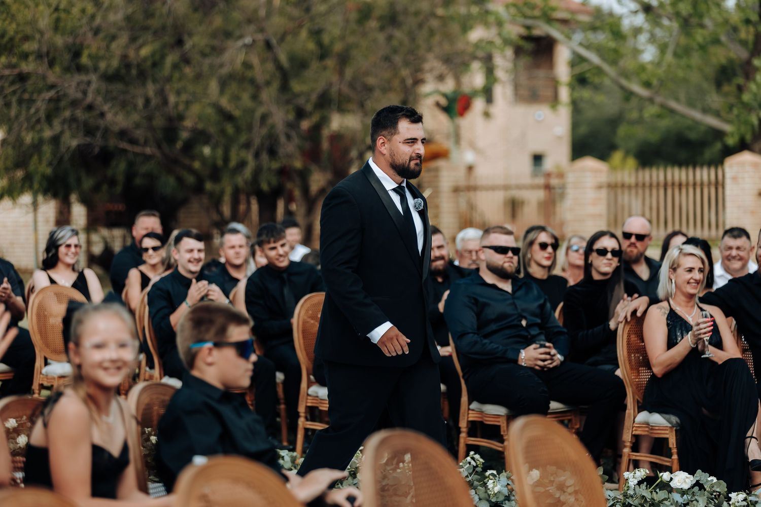 Wedding guests gather outdoors for a celebration ceremony in a garden setting with natural lighting.