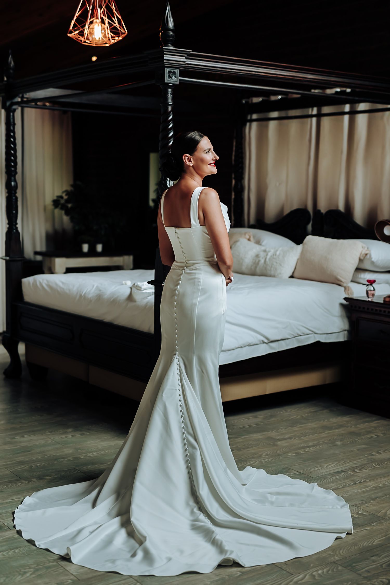 Back view of a bride in an elegant white wedding dress with button details and a flowing train on dark hardwood floors.