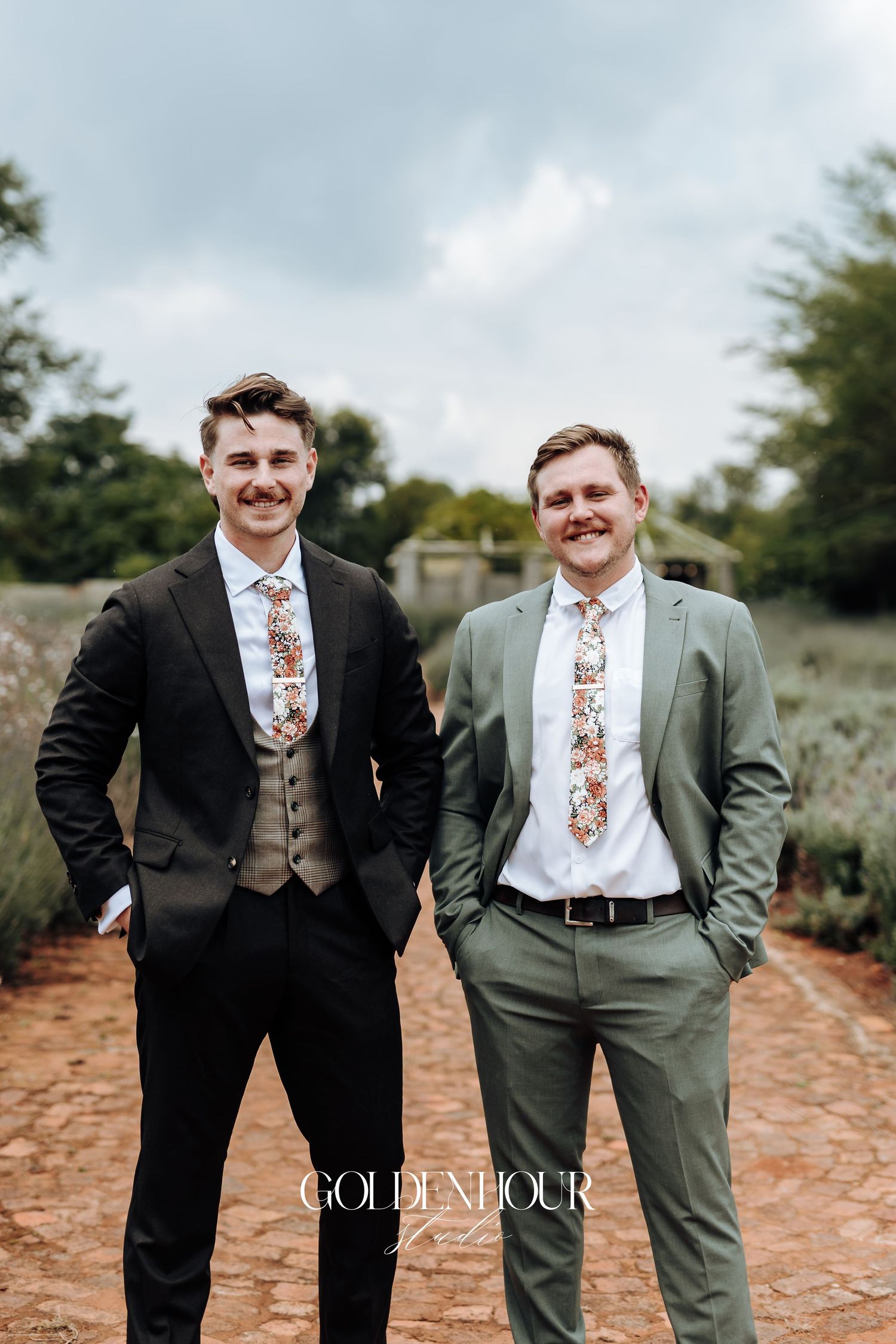 Two groomsmen in contrasting black and olive suits pose together in an outdoor setting.