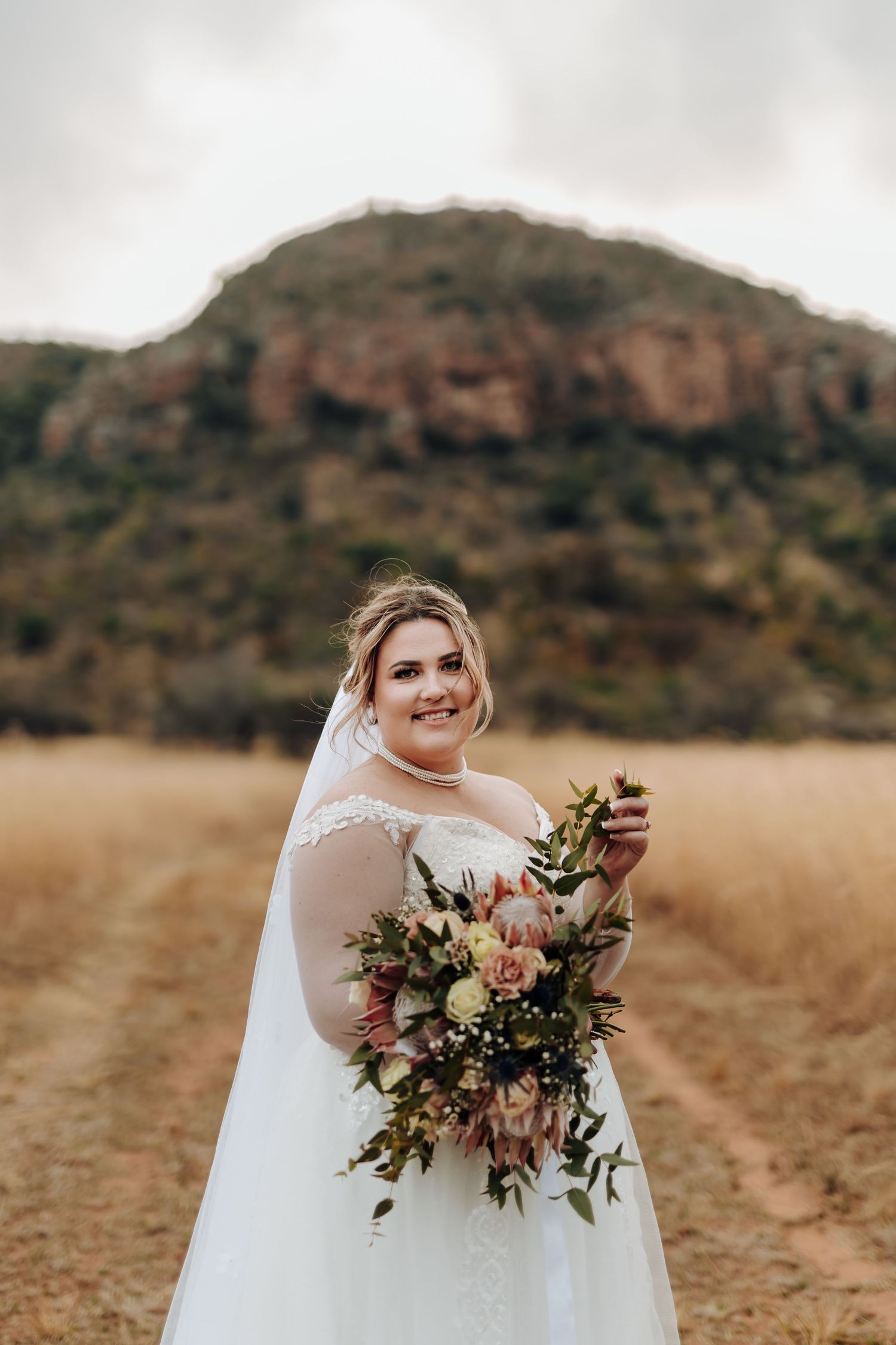 A bride in a white dress holds a stunning autumn bouquet of pink and burgundy flowers against a mountain backdrop.