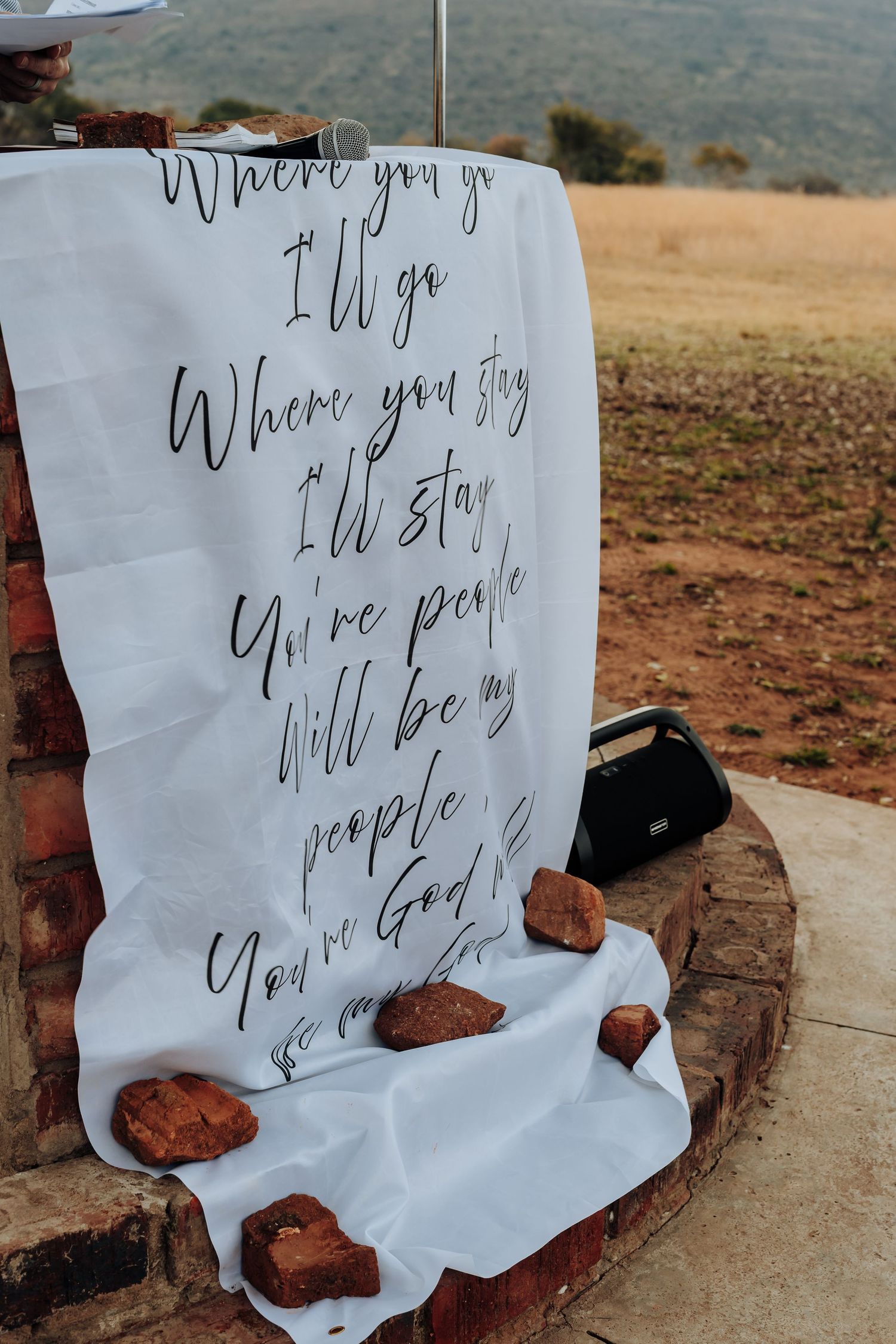 White fabric with handwritten wedding vows and song lyrics displayed outdoors against a rural mountain landscape with red rocks.