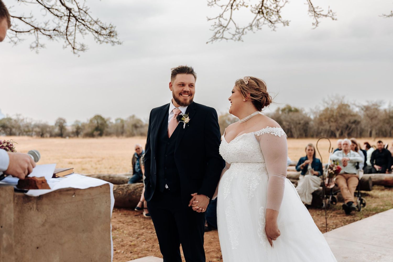 A couple shares a joyful moment during their outdoor rustic wedding ceremony in a field on a cloudy day.