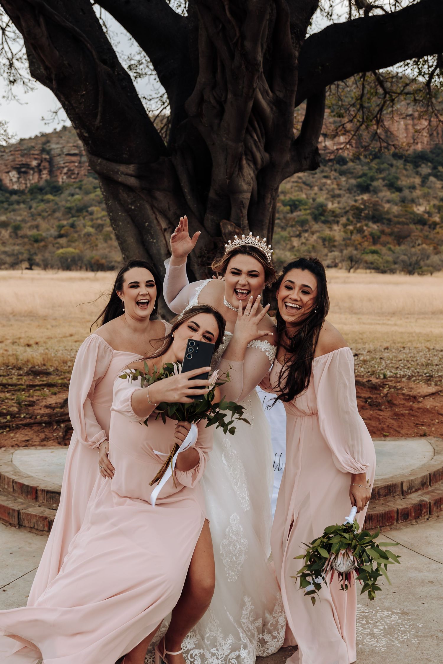 A group in pale pink and white dresses laughing joyfully together under a large tree in a desert landscape.