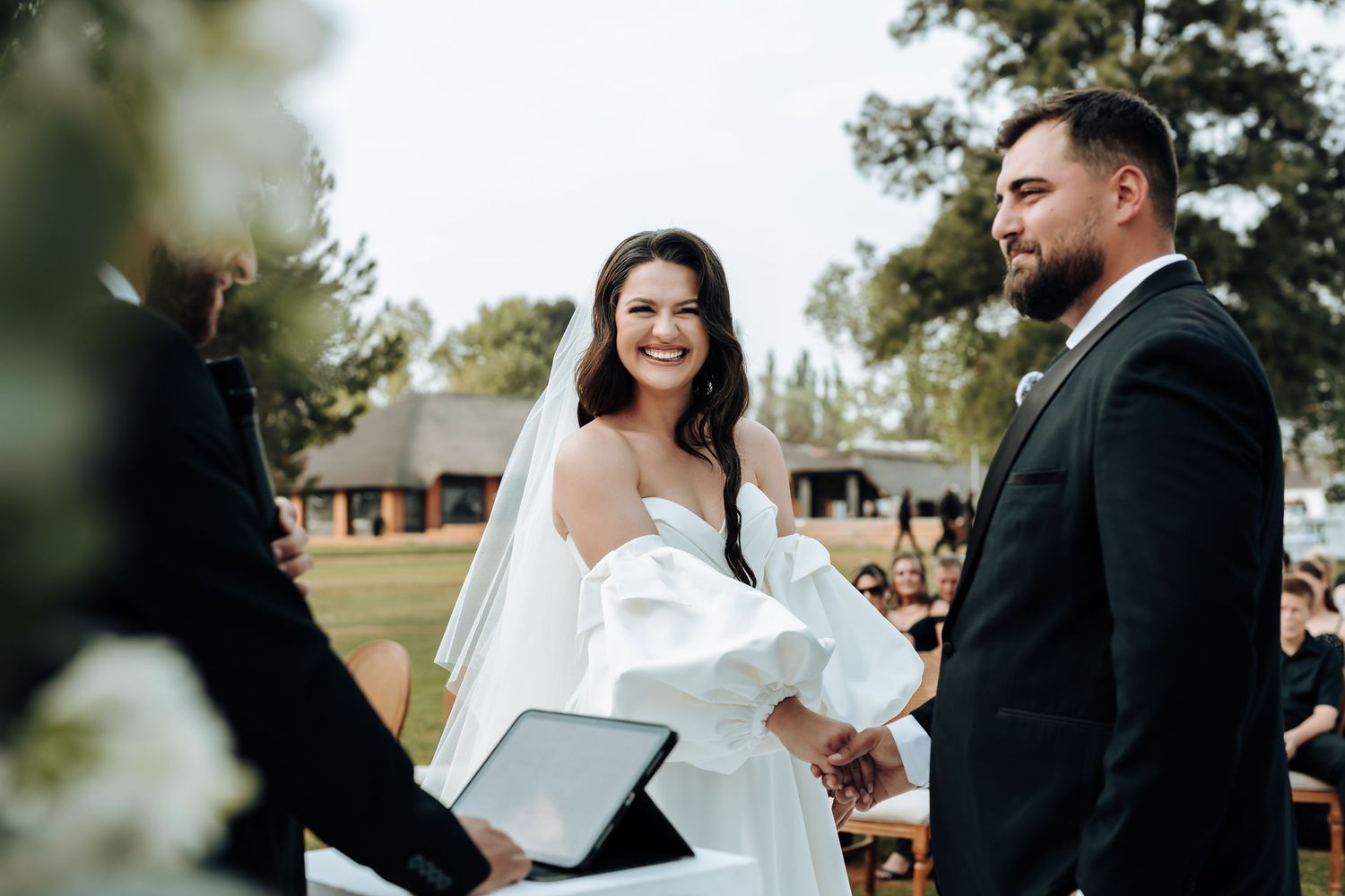 Outdoor wedding ceremony with couple exchanging vows under a beautiful tree canopy on a sunny day.