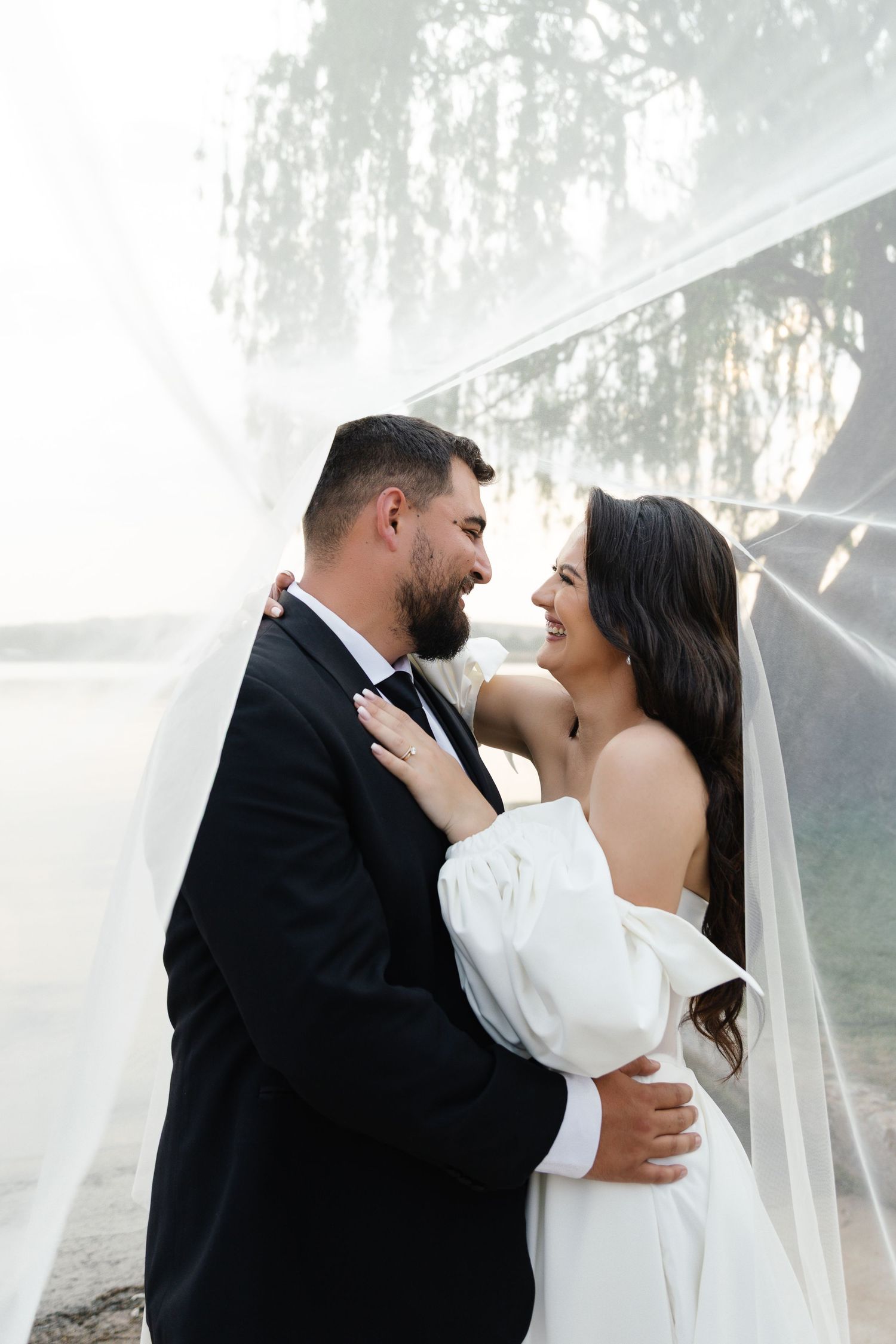 A romantic wedding couple embraces under a misty forest canopy with flowing veil and elegant attire.