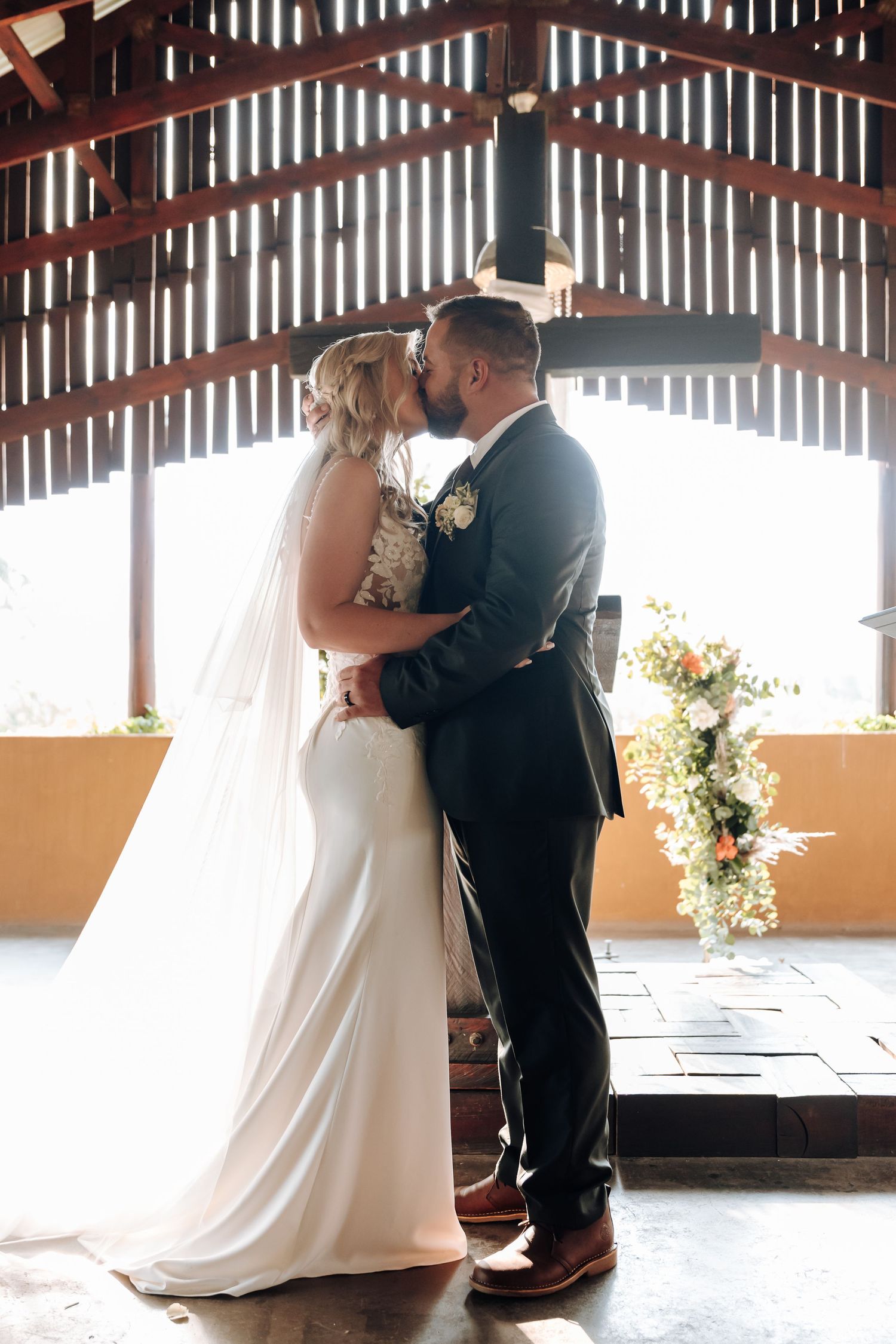 A romantic wedding couple shares a kiss under a rustic wooden beam pavilion with floral decorations.