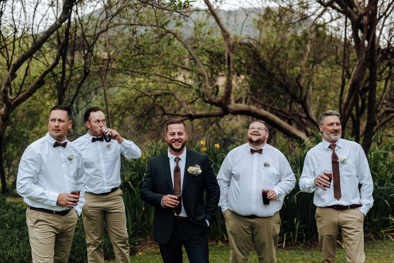 A group of groomsmen in suits and bowties laugh together while holding drinks during an outdoor wedding celebration.