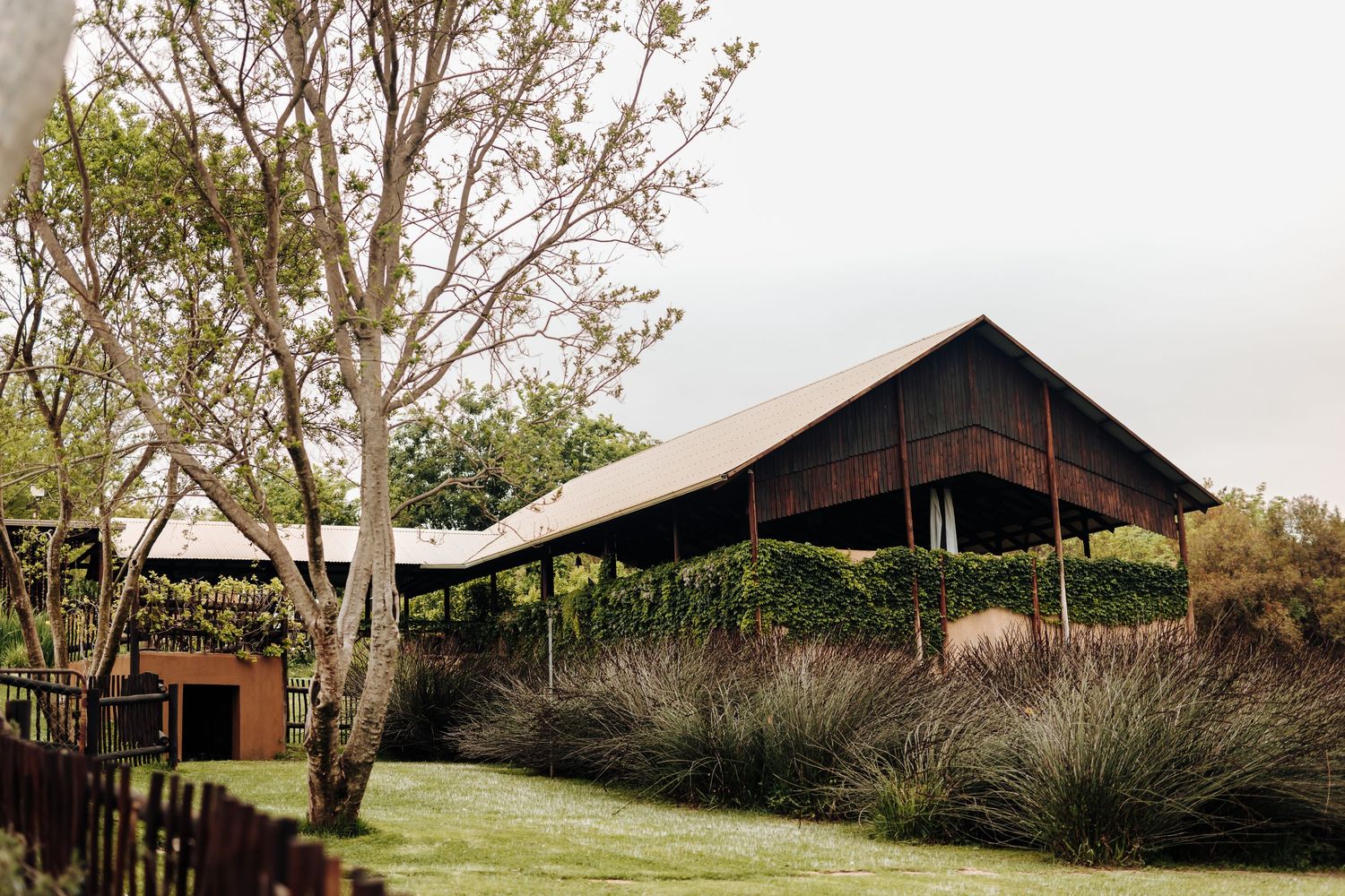 A rustic wooden barn with dark timber exterior stands against a cloudy sky surrounded by leafy trees and natural landscaping.