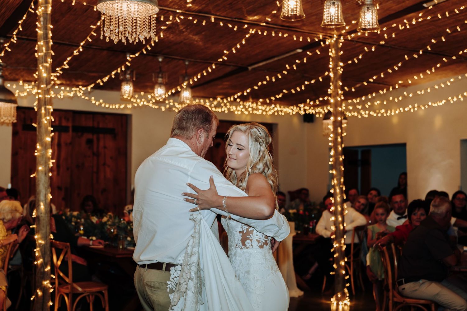 Wedding party dancing and celebrating under warm string lights at an indoor reception venue.