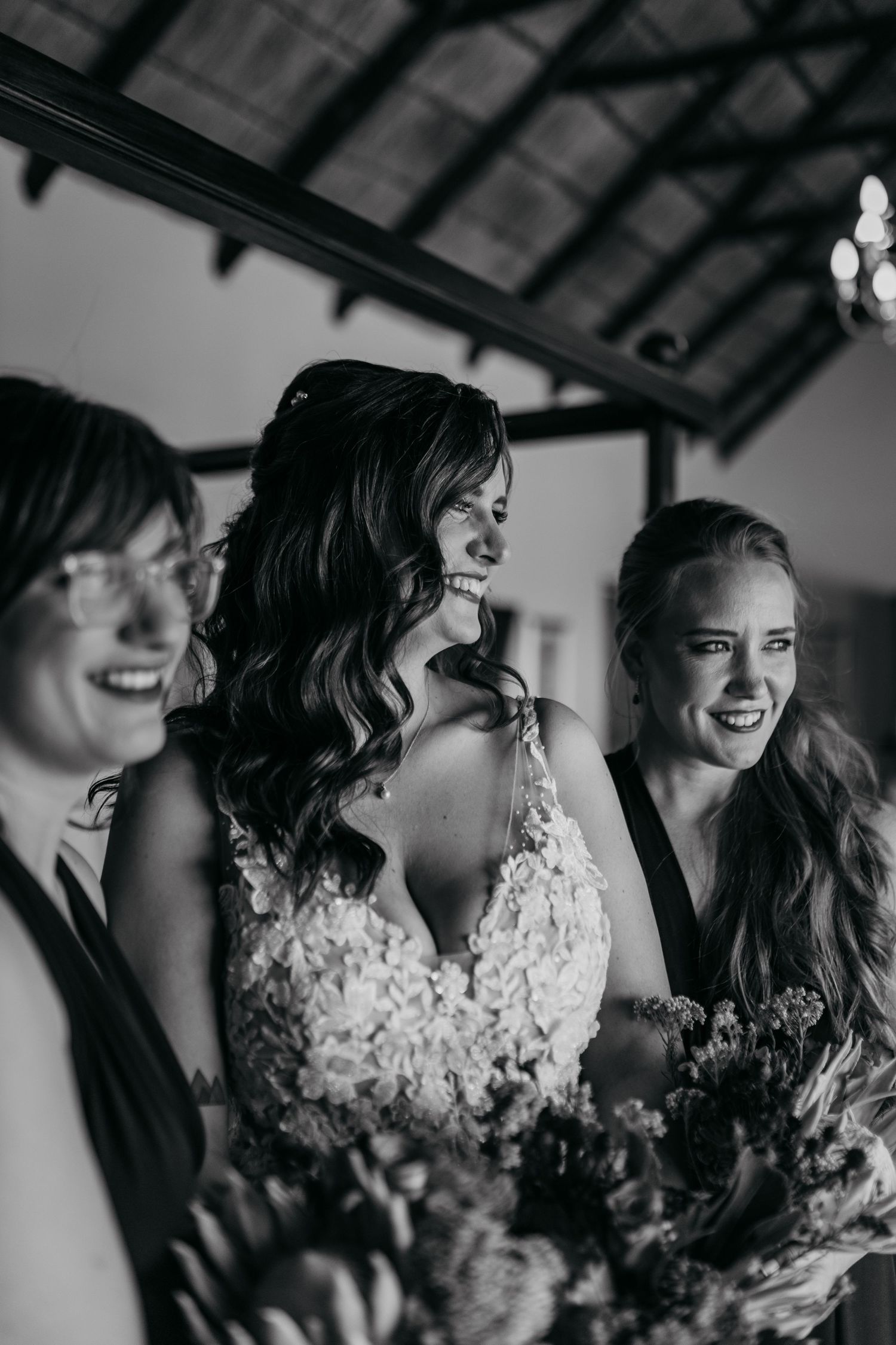 Black and white candid photo of three people laughing and smiling together while holding bouquets at a wedding.
