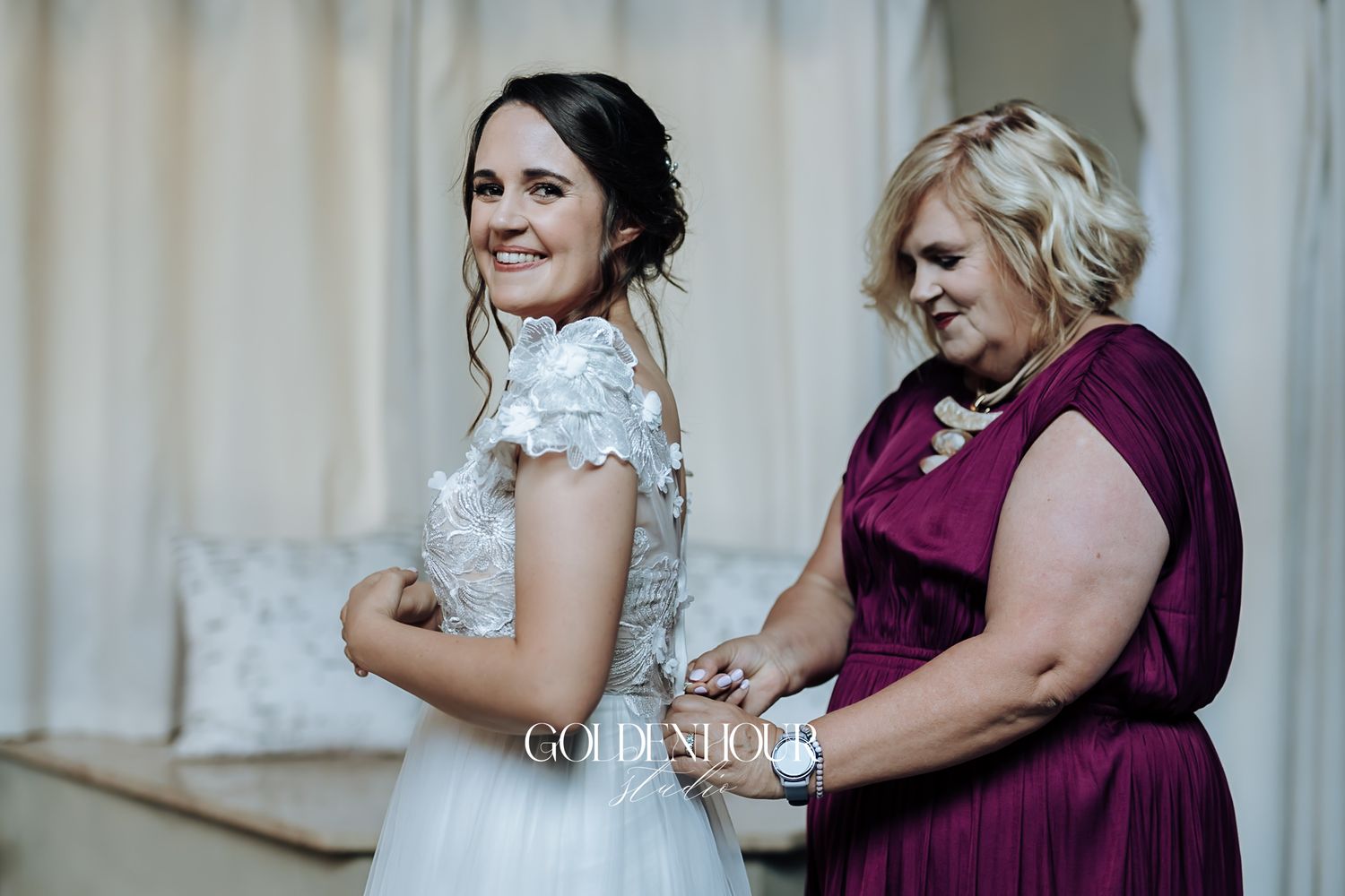 Bride in lace wedding dress being helped by woman in burgundy dress during bridal preparations.