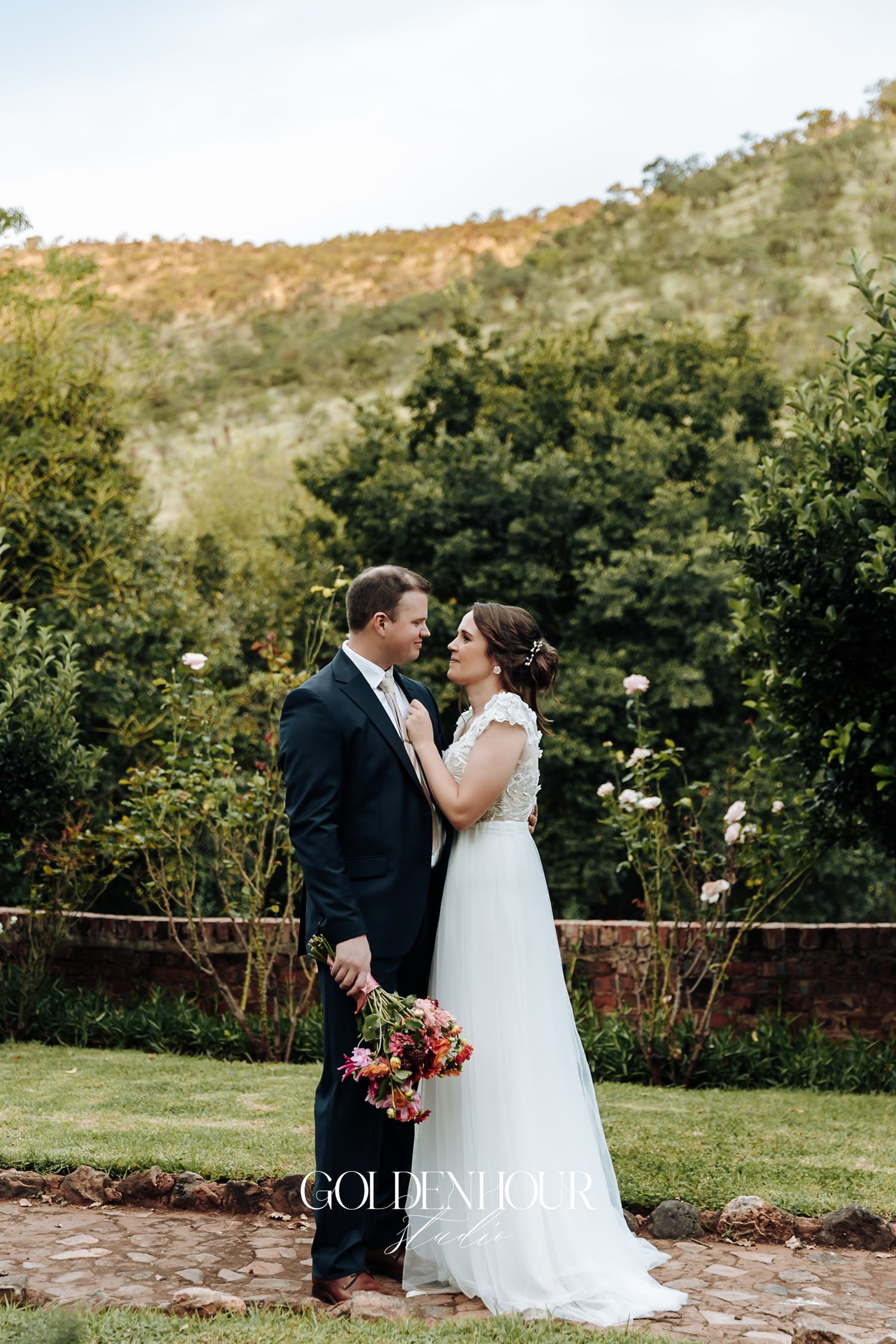 Bride and groom gazing at each other in lush garden with scenic mountain backdrop behind them.