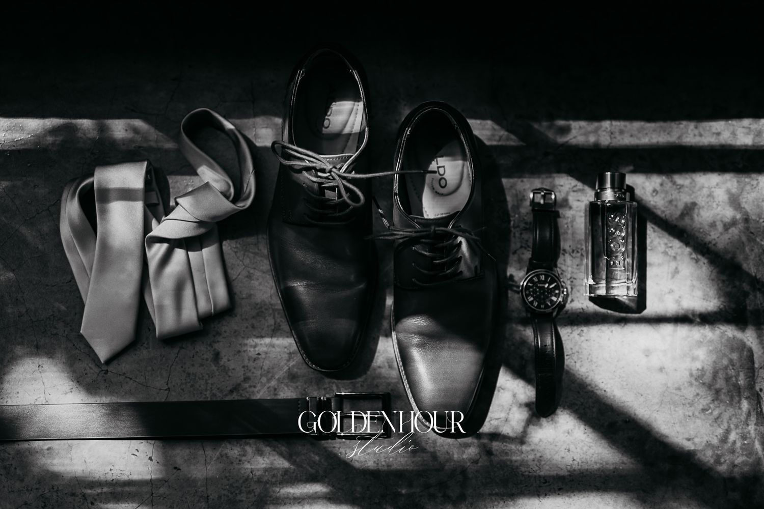 Black and white flat lay of mens wedding accessories including dress shoes, tie, watch, and cologne bottle.