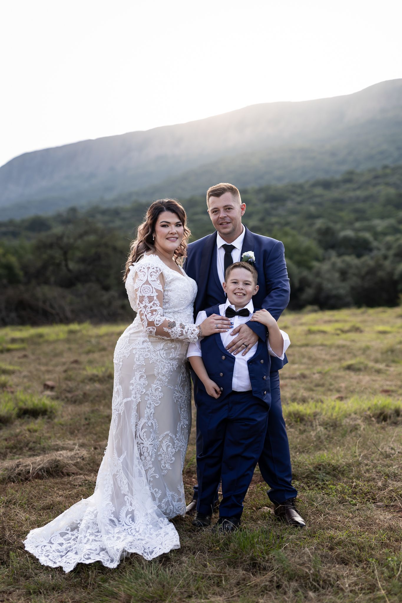 Wedding couple poses with young ring bearer in matching navy suits against mountain landscape.