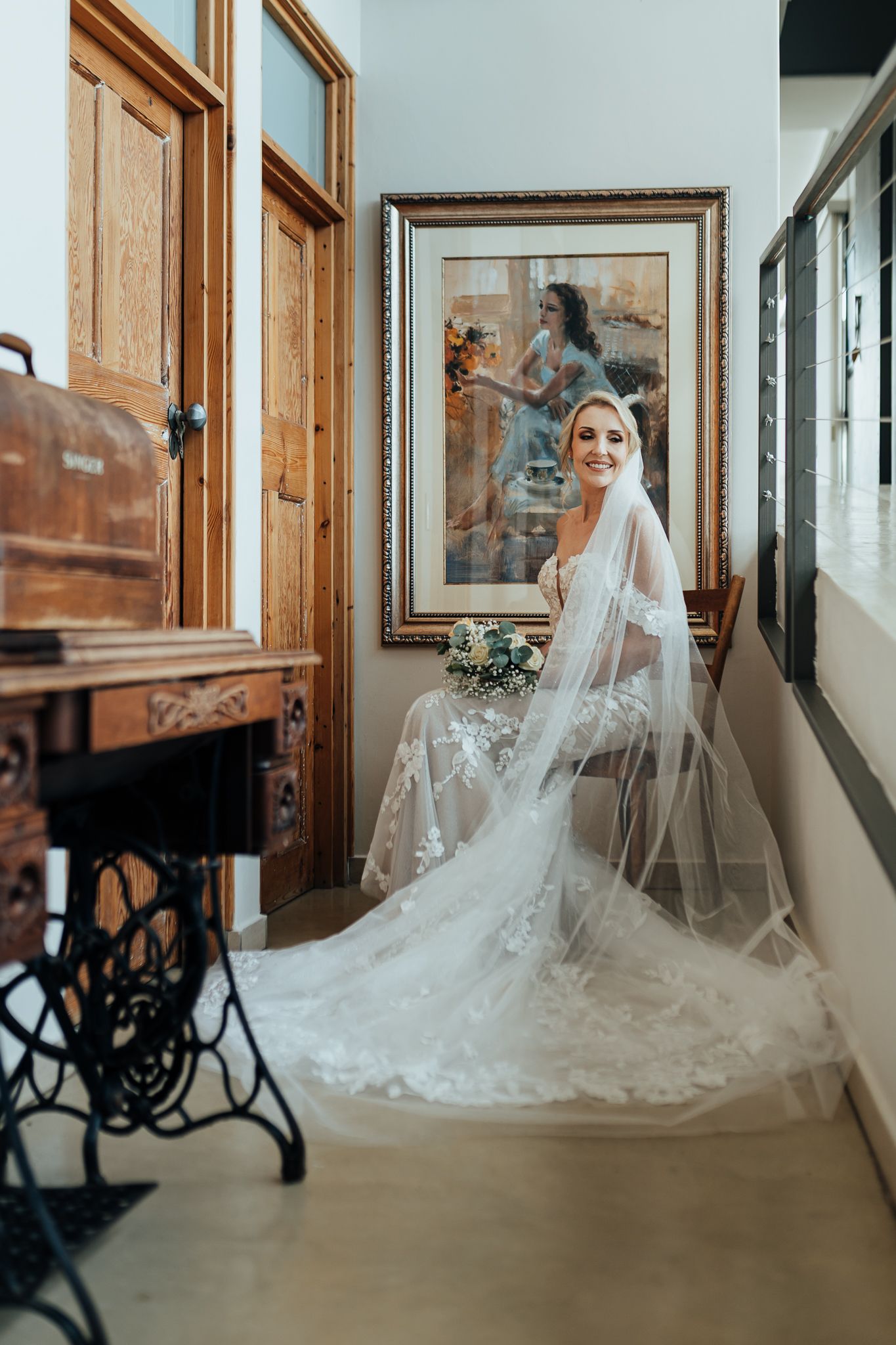 Bride in lace gown and veil poses by vintage sewing machine and framed artwork.