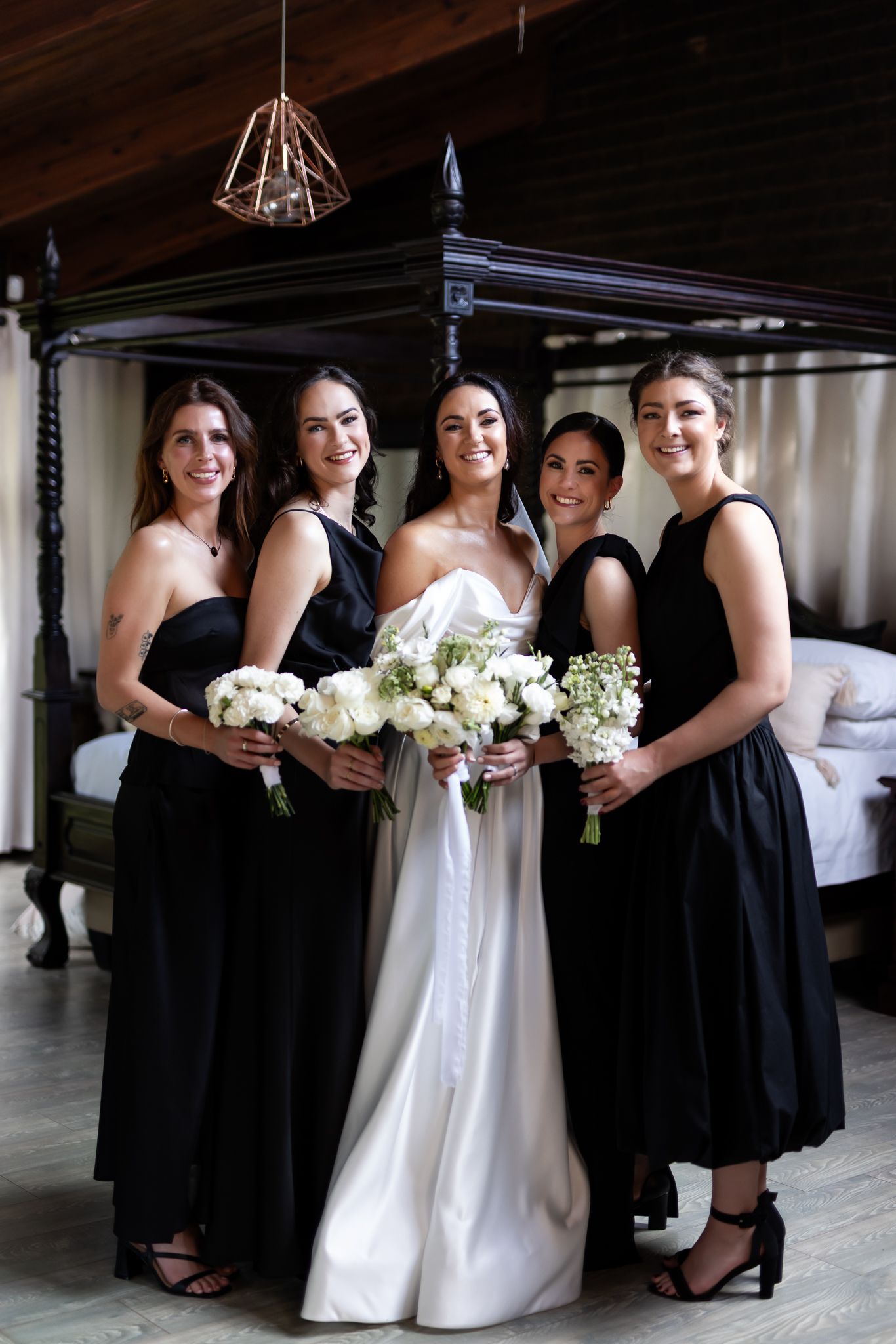 Bride in white gown poses with bridesmaids in black dresses holding white floral bouquets.