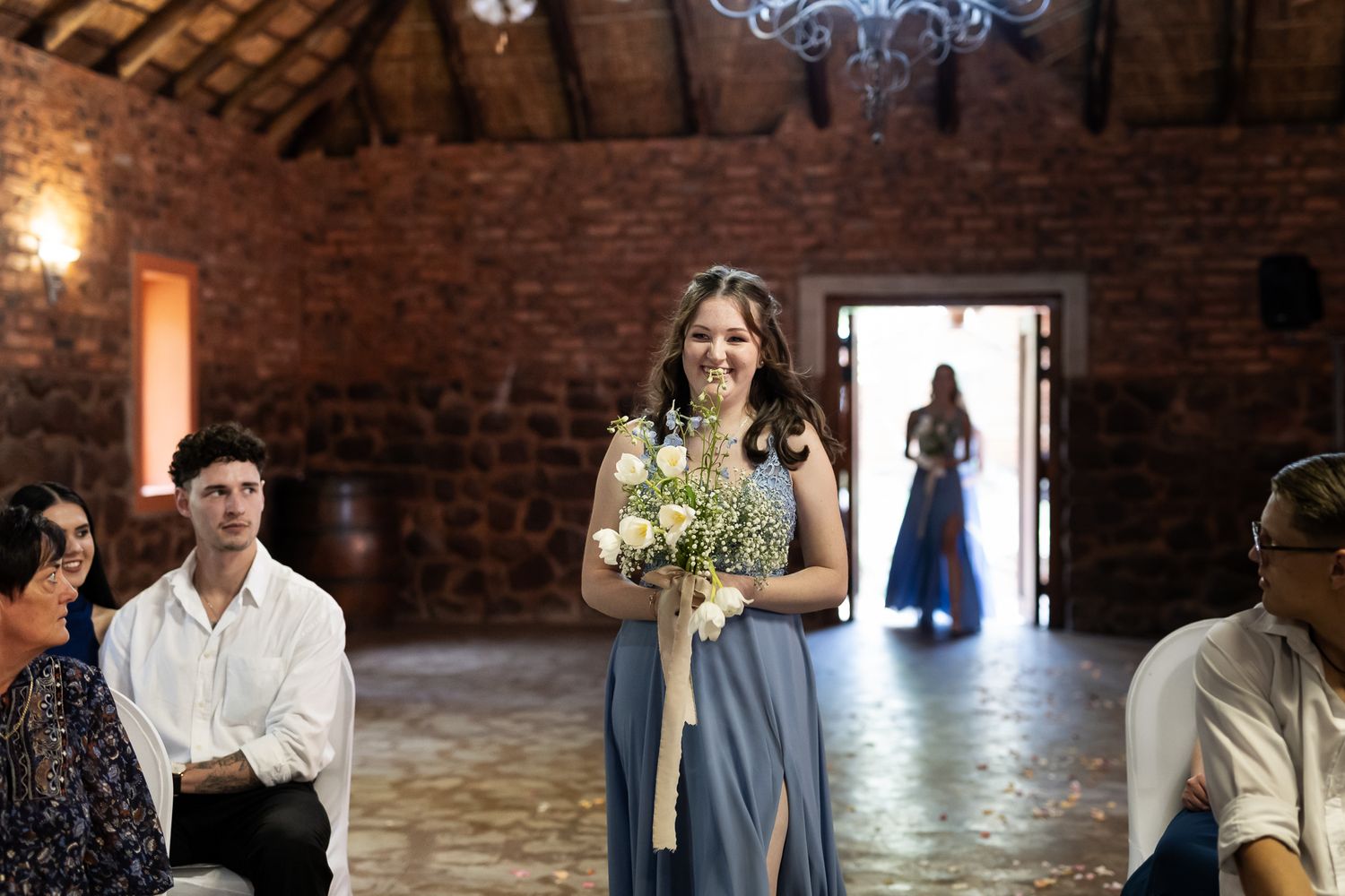 Elegant bridesmaid in light blue dress holds bouquet inside rustic brick venue with exposed beams and chandelier.