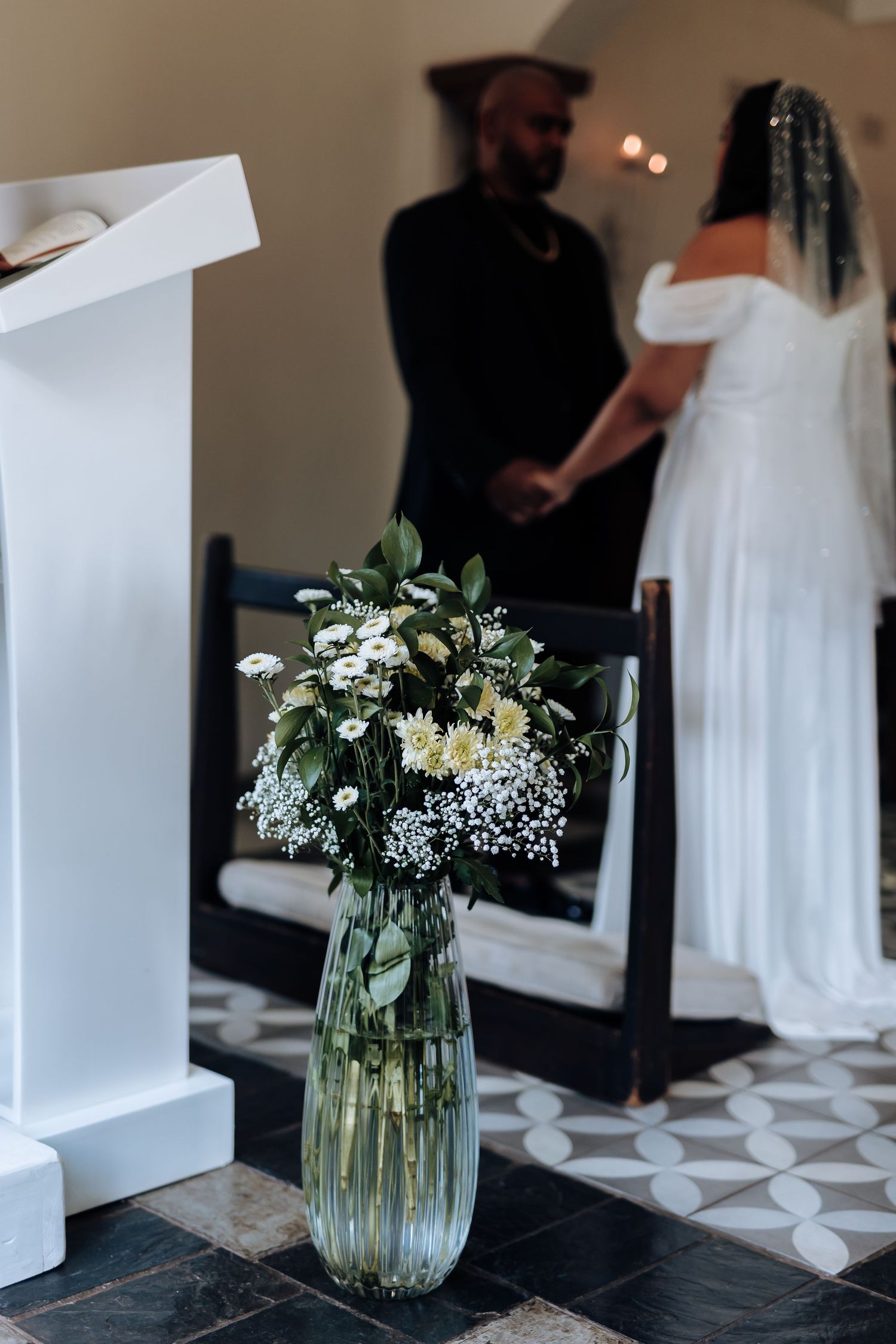 A floral arrangement in a glass vase with white flowers and greenery during a wedding ceremony.