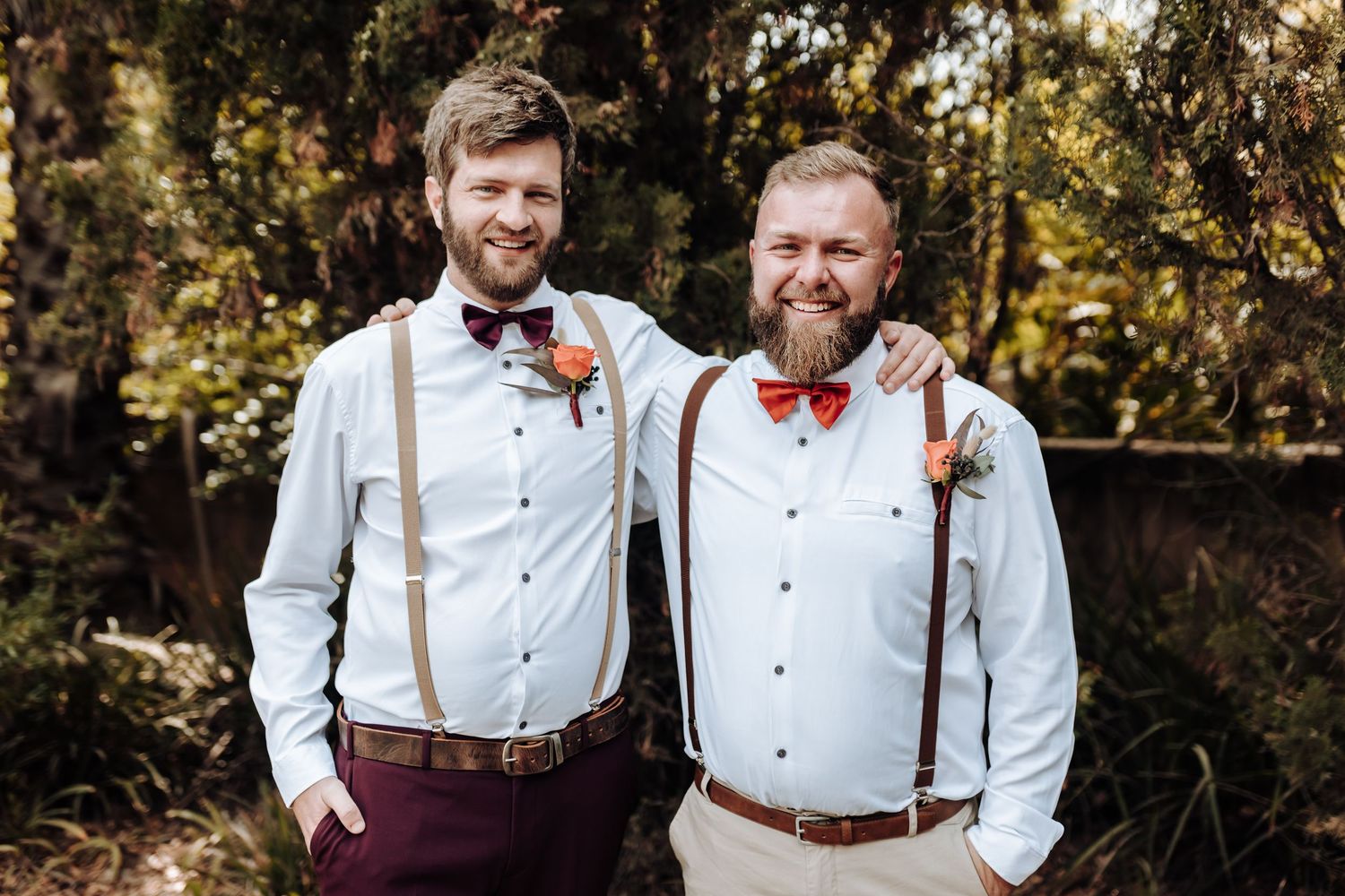 Groomsmen in coordinated white shirts, suspenders and bow ties pose together against trees.