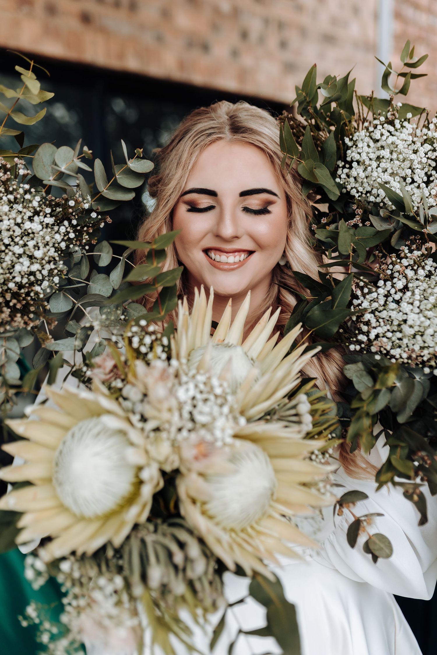 A bride smiles joyfully surrounded by eucalyptus greenery and white protea flowers in her wedding bouquet.
