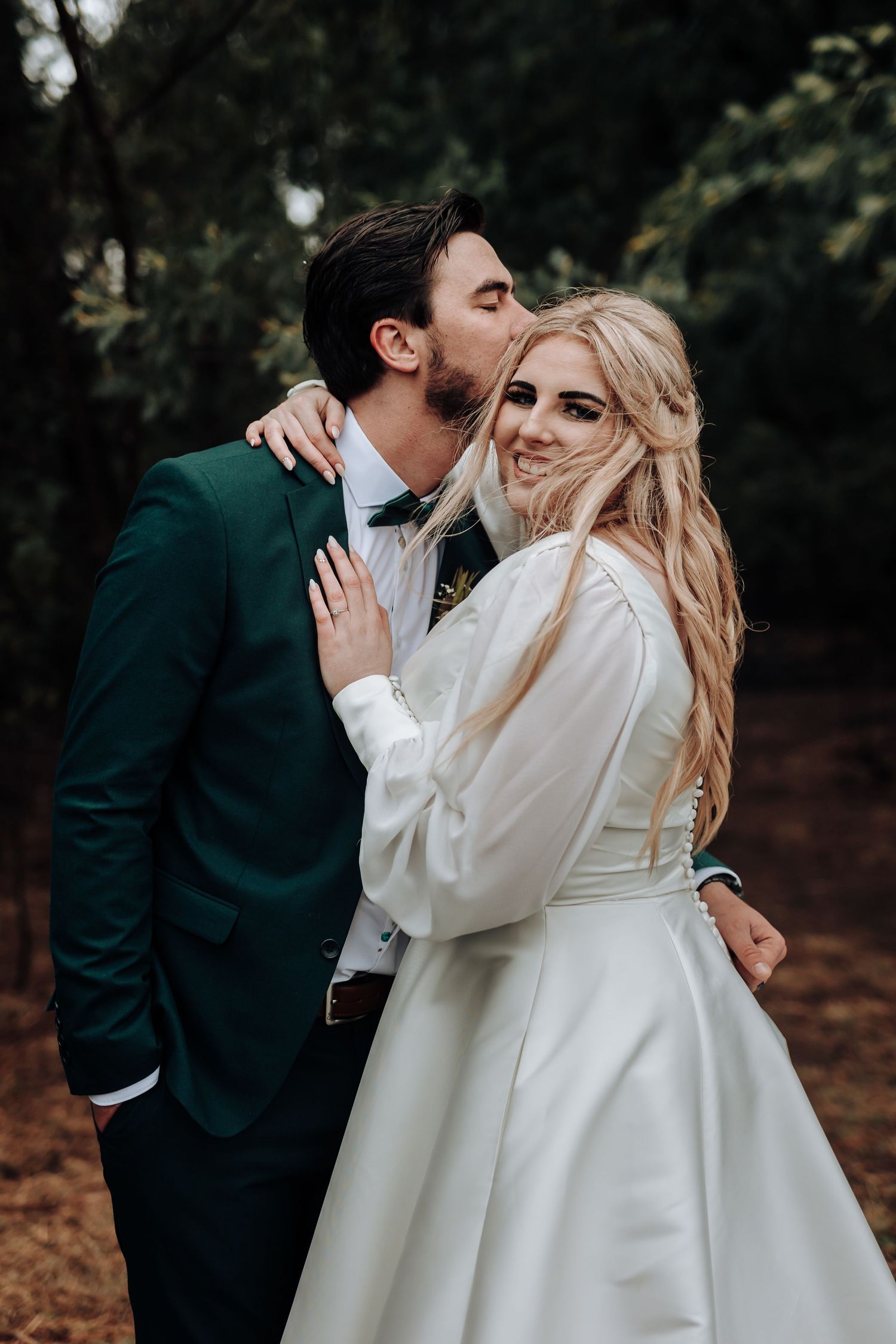 A romantic wedding photo of a couple embracing outdoors, with the groom in emerald green and bride in white gown.