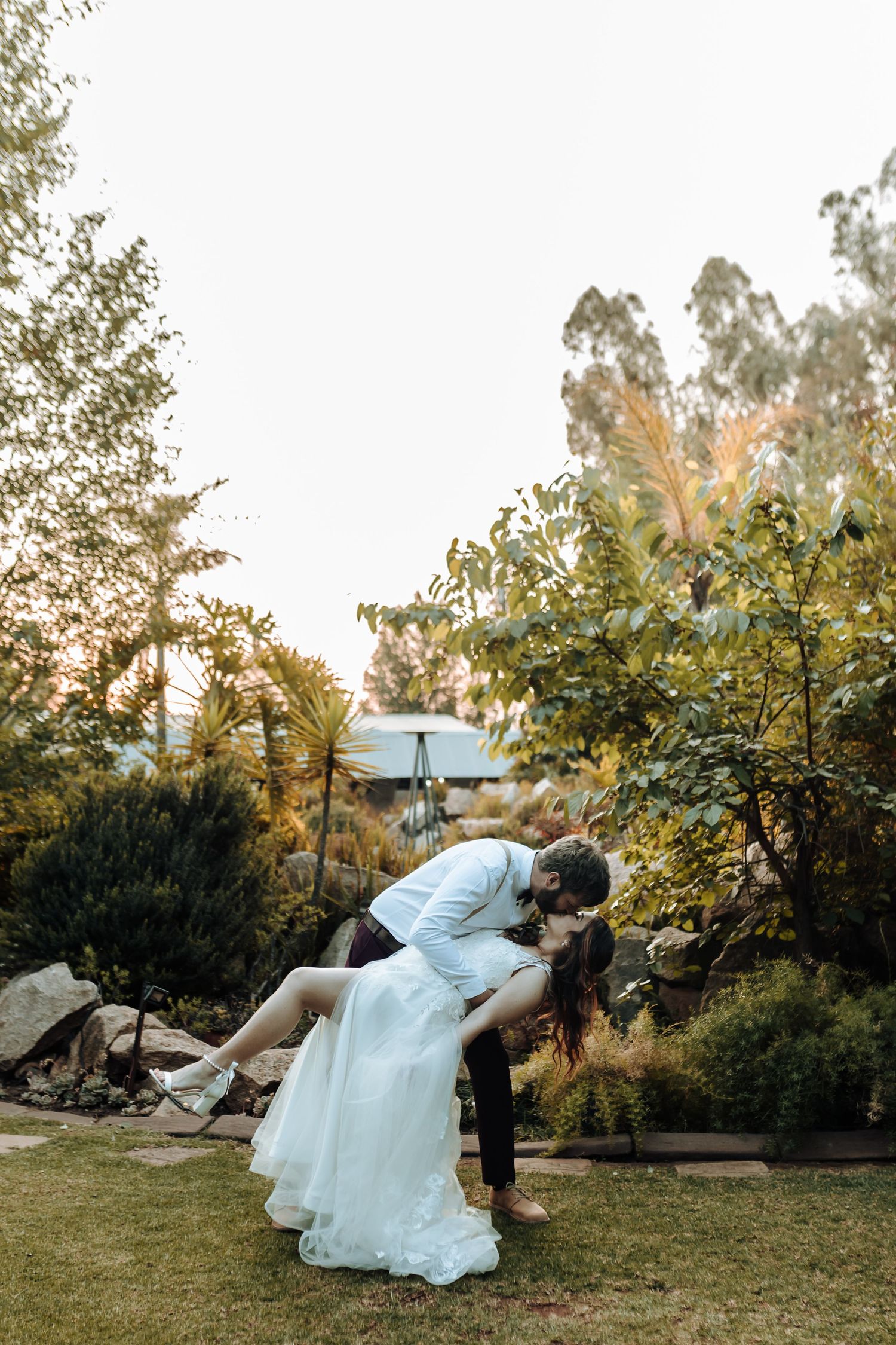 A romantic dip pose outdoors with sunlight filtering through trees in a garden wedding setting.