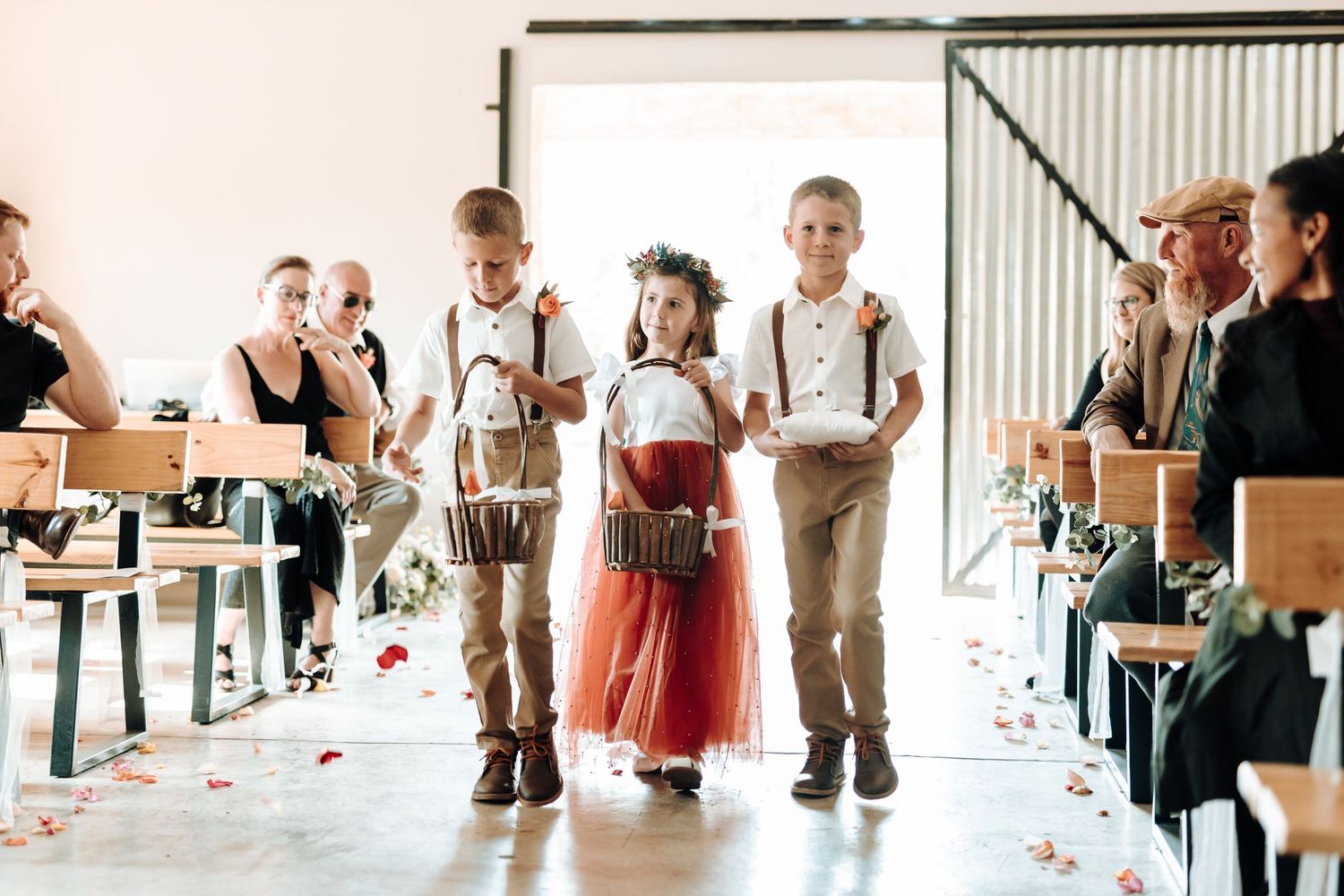 Children walk down aisle at wedding ceremony while guests watch from wooden benches.