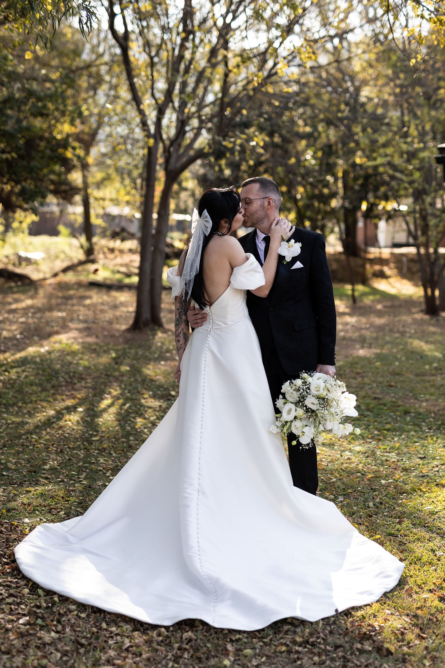 A romantic wedding couple embraces in a sunlit wooded area, the bride wearing an elegant white gown with a flowing train.