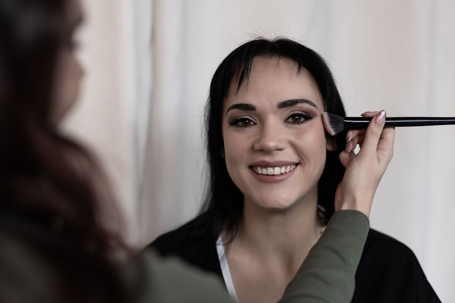 A smiling woman applies makeup in the mirror while wearing a dark green top.