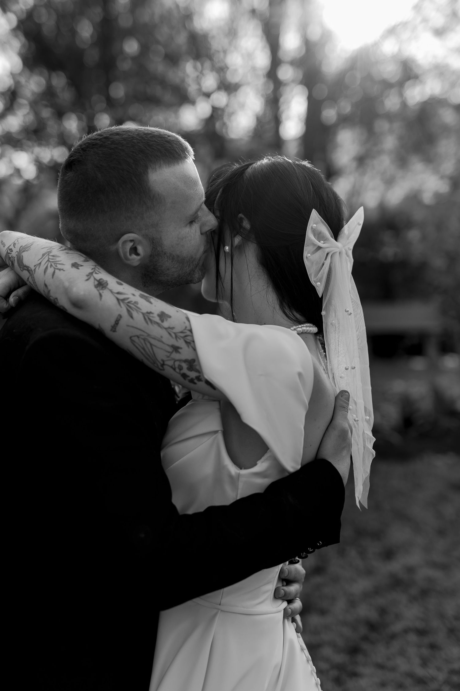 Black and white romantic photo of an intimate embrace between two people in wedding attire outdoors with soft bokeh lighting.