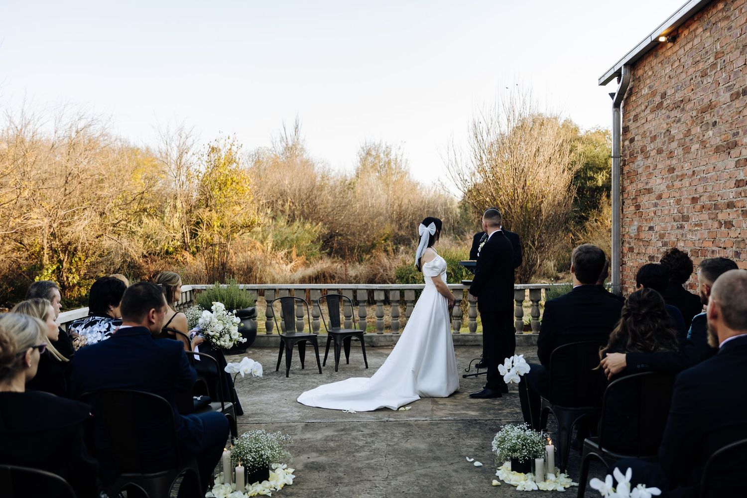 An outdoor wedding ceremony takes place on a stone terrace with autumn foliage in the background and floral arrangements.
