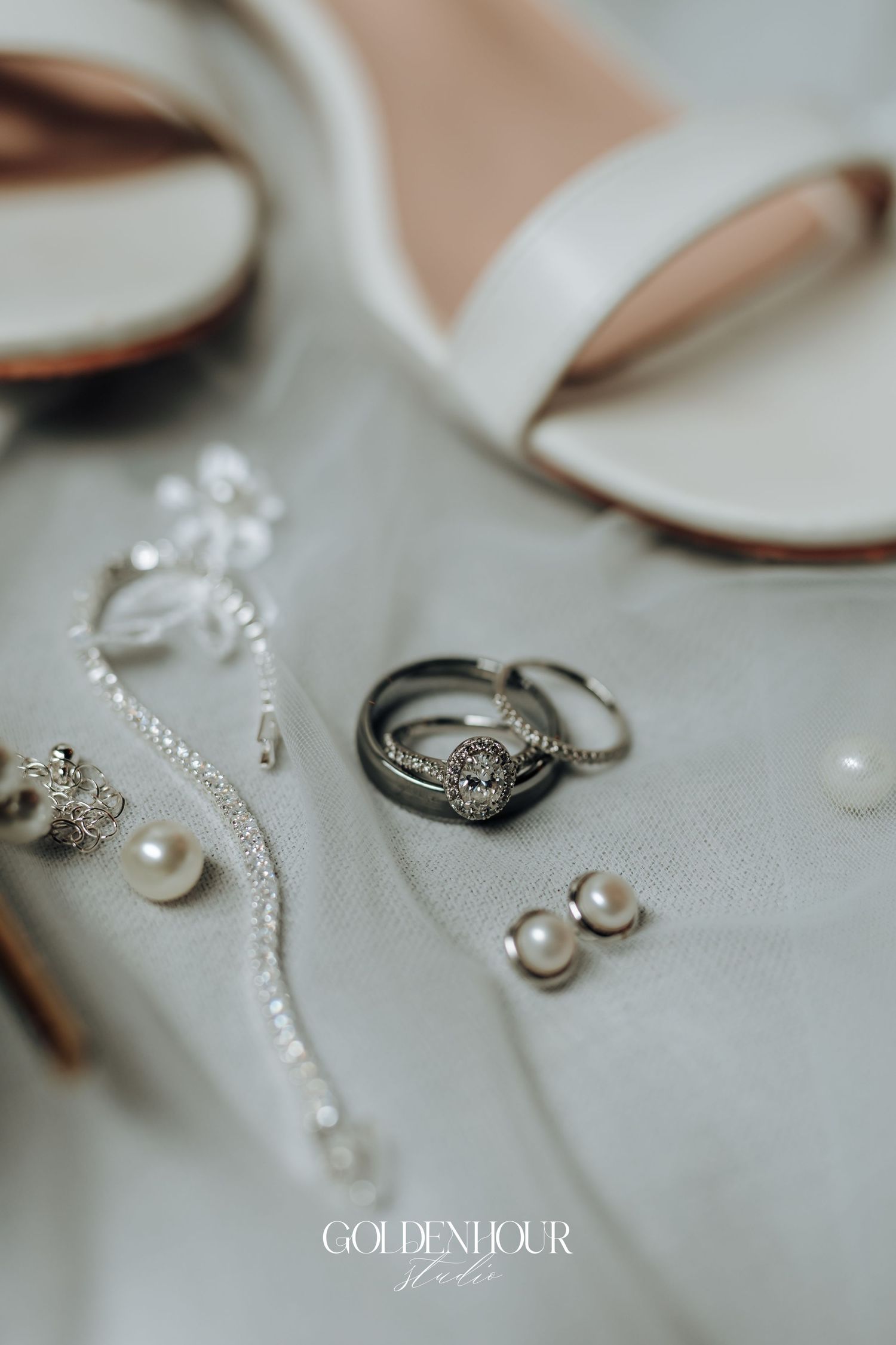 Elegant bridal accessories flat lay featuring diamond rings, pearl earrings, crystal jewelry, and white heeled sandals on soft fabric.