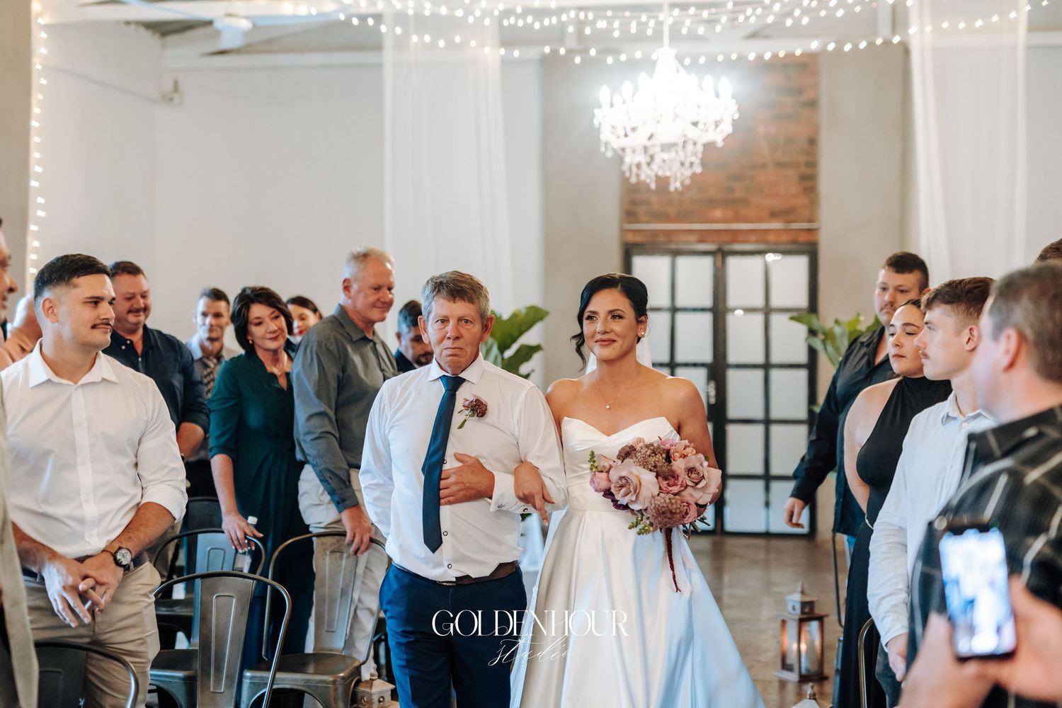 Smiling bride in strapless gown walks down aisle with escort under fairy lights at indoor wedding ceremony.