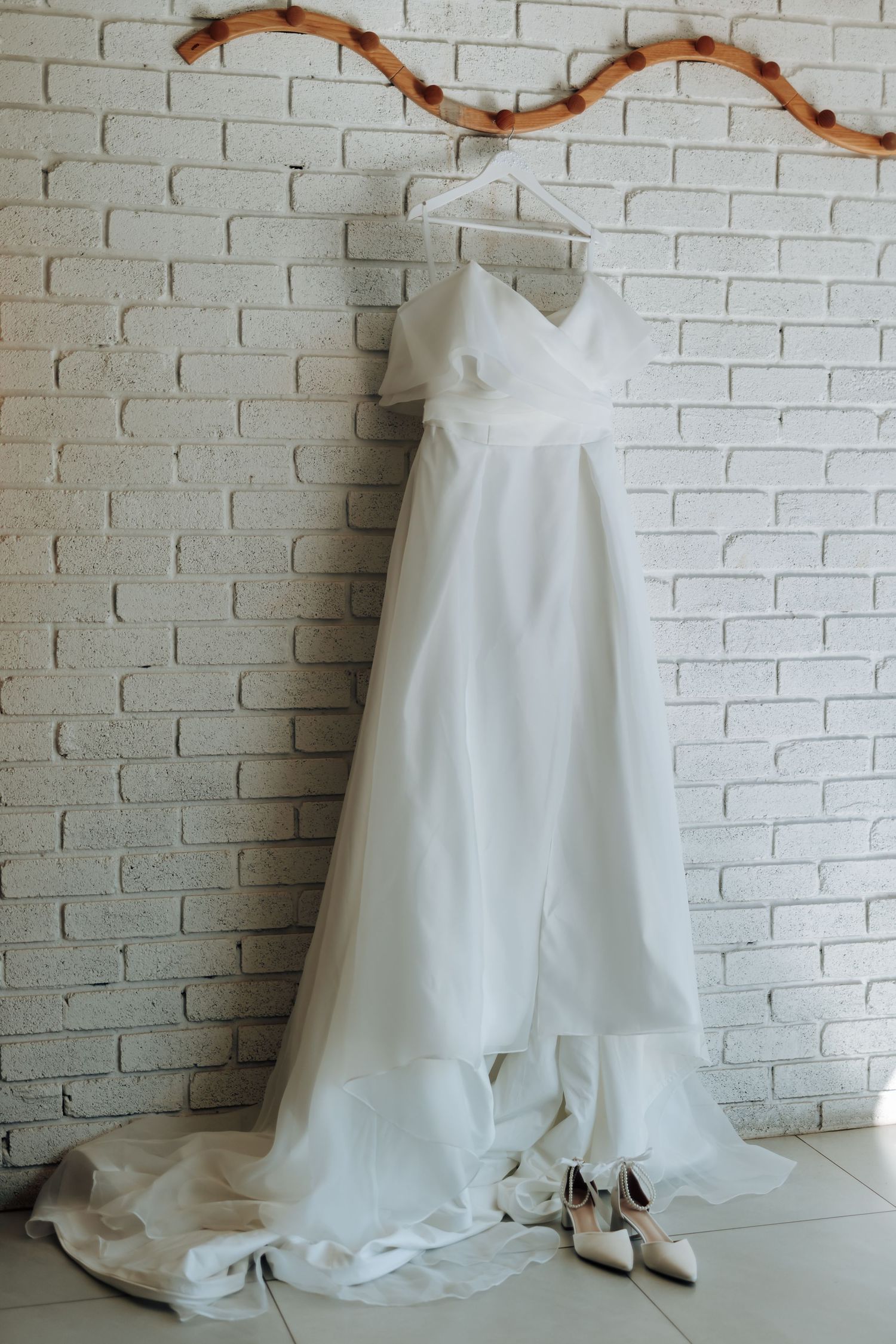 Elegant white wedding dress hanging on a rustic branch against a brick wall, with bridal heels below.