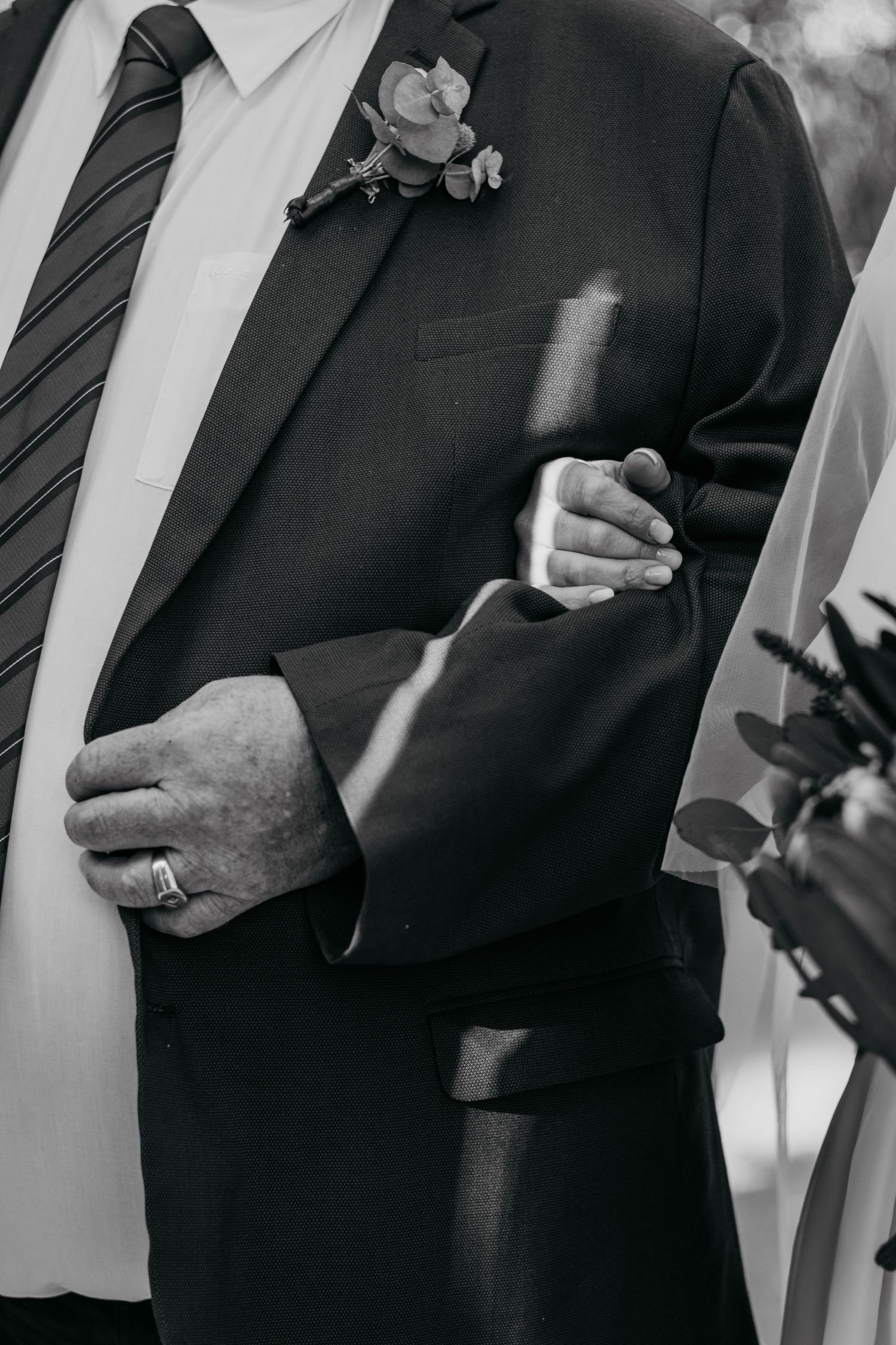 Black and white photo of groom in suit with boutonniere, holding hands with bride at wedding ceremony.