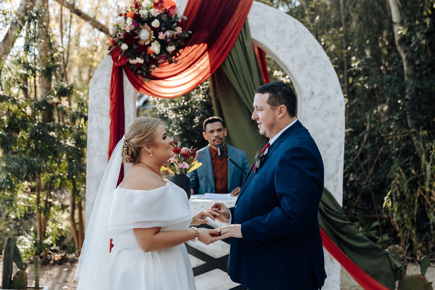Couple exchanging vows at outdoor wedding ceremony with red and white floral arch and officiant.