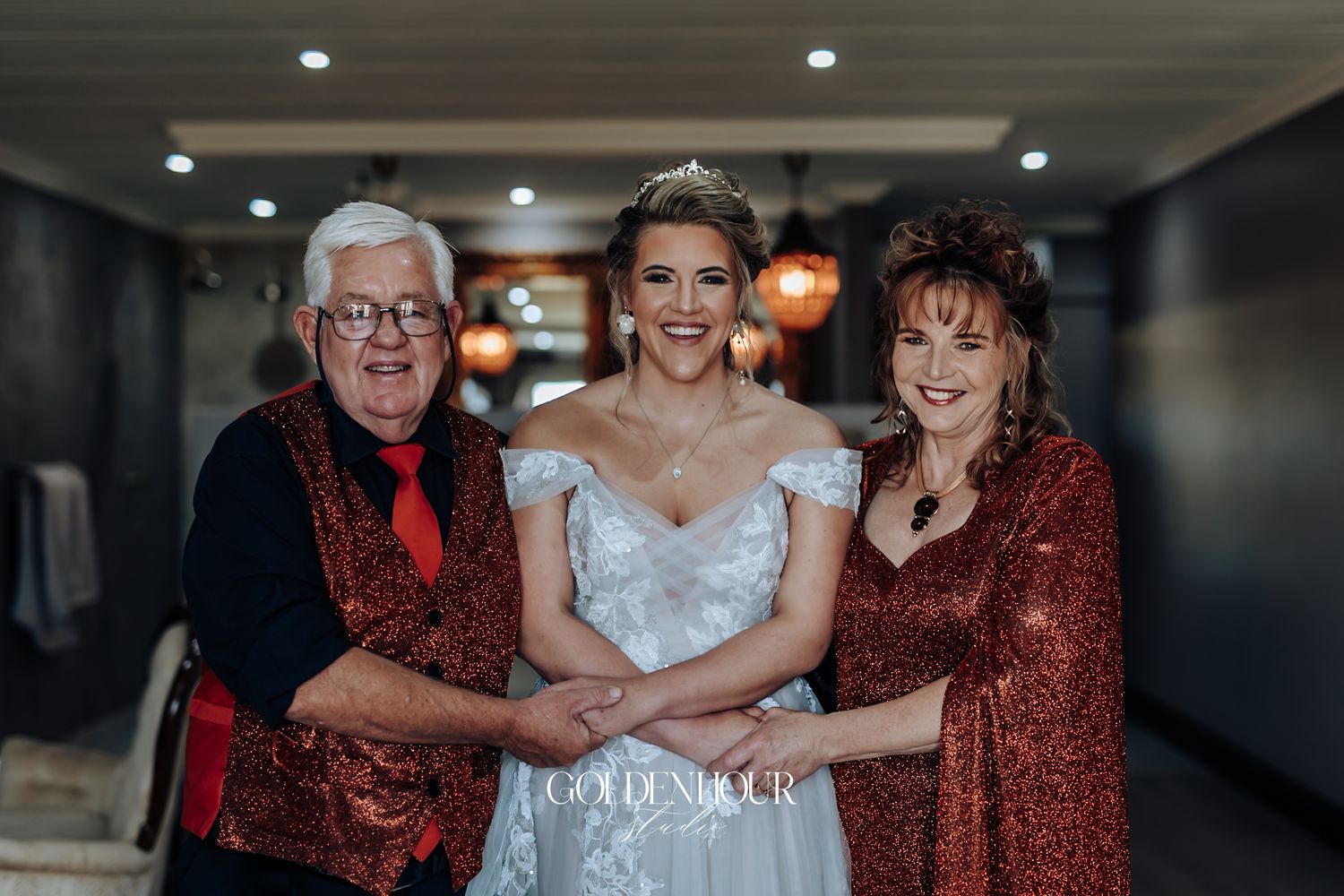 Bride in white off-shoulder gown posing with parents in red sequin outfits at wedding.