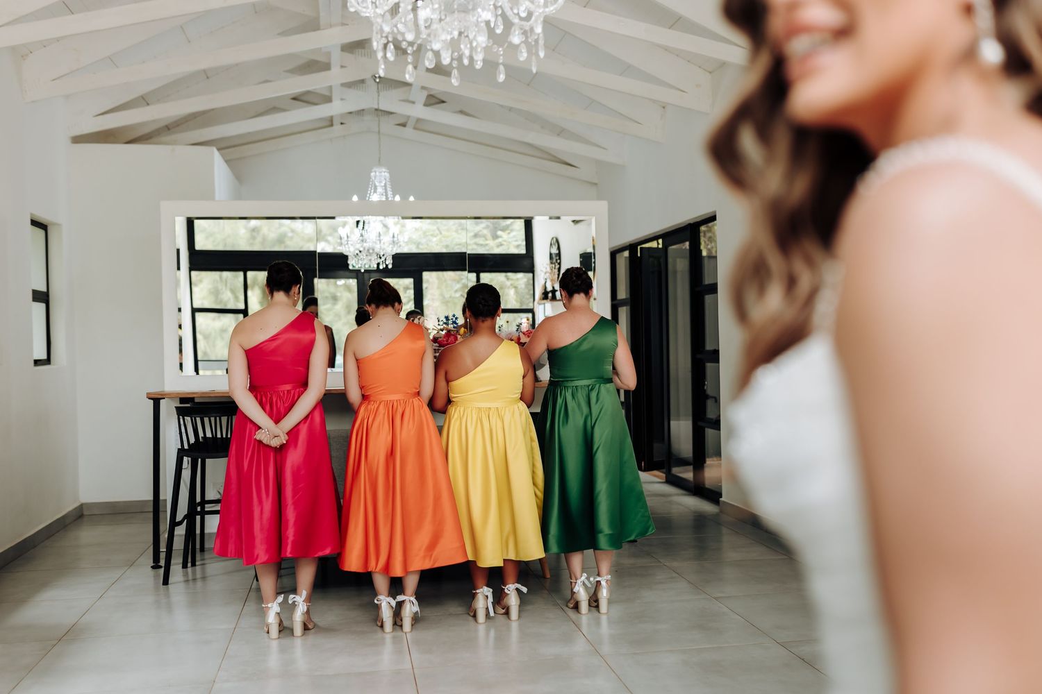 Bridesmaids in vibrant red, orange, yellow and green dresses stand in a row inside an elegant white room with chandelier.