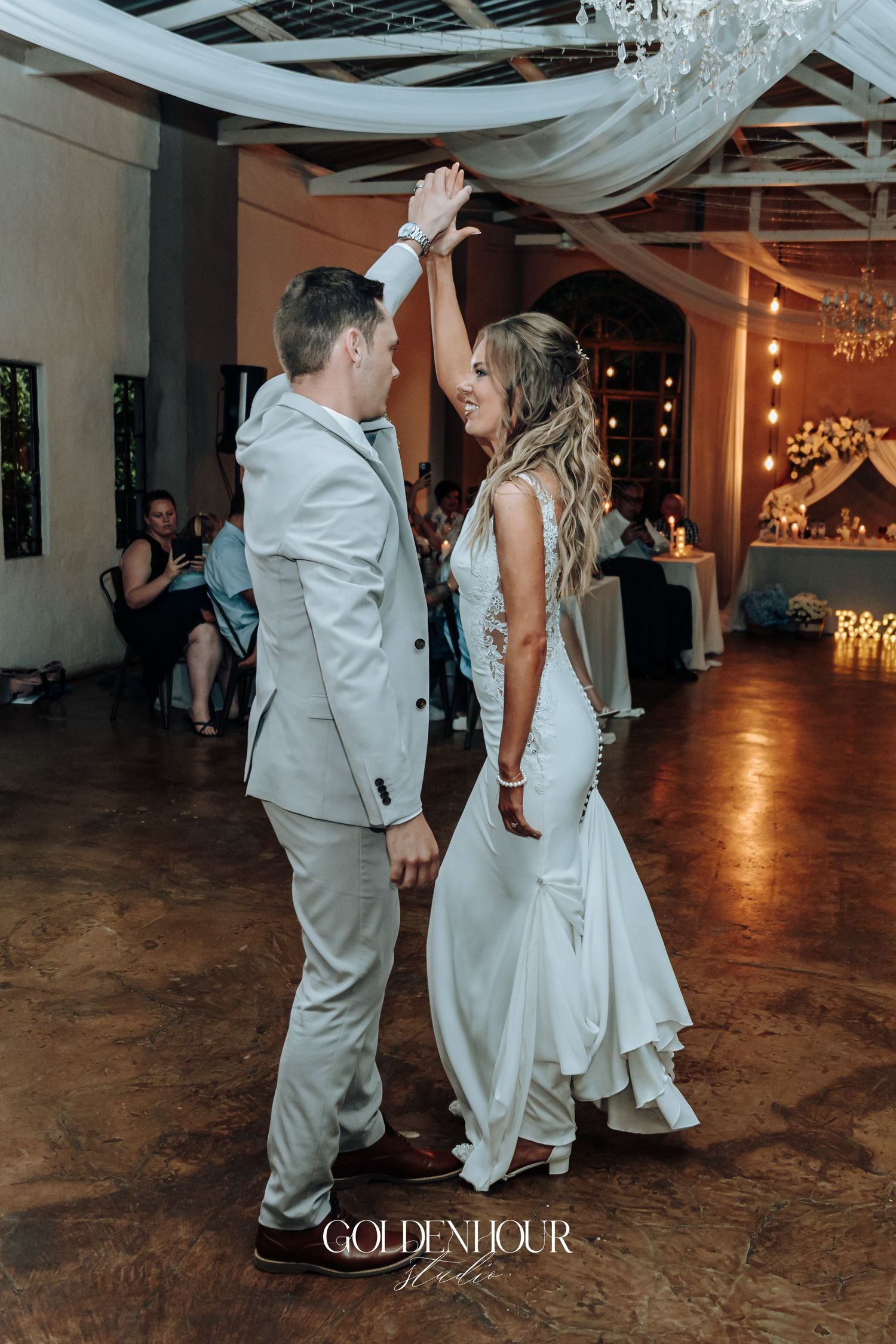 Newlyweds share their first dance on a rustic wooden floor under romantic lighting and draped fabric.