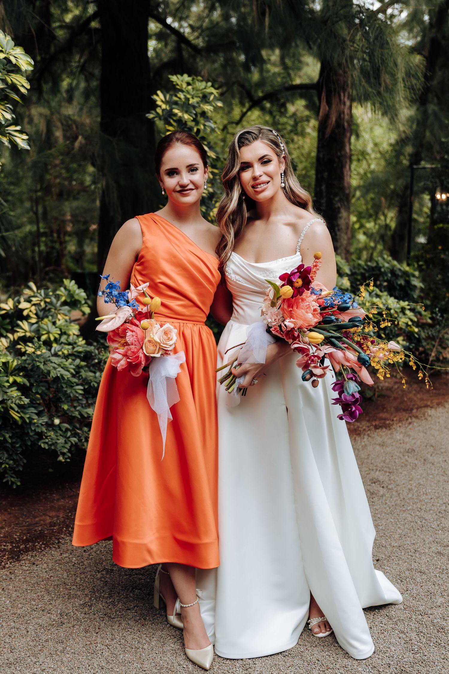 Two people in orange and white dresses hold colorful floral bouquets while posing together in a garden setting.