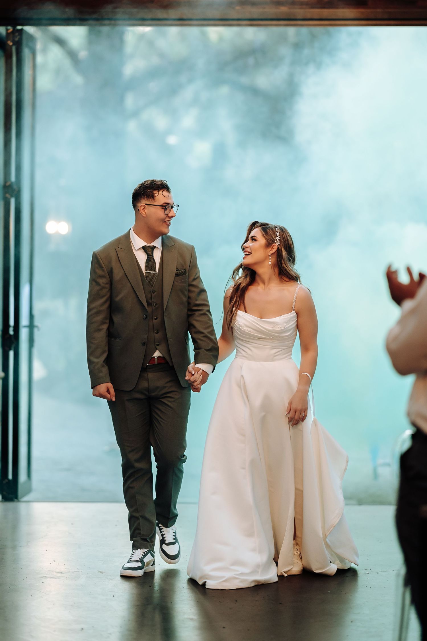 A couple in wedding attire walk together indoors, with the groom wearing sneakers and a grey suit.