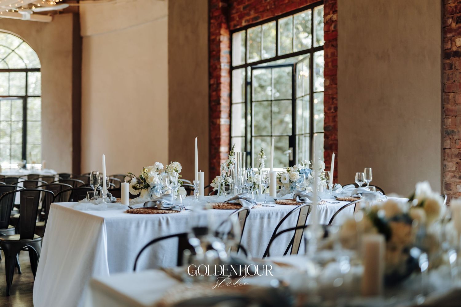 Elegant wedding reception venue with round tables, white linens and arched windows in a rustic brick room.