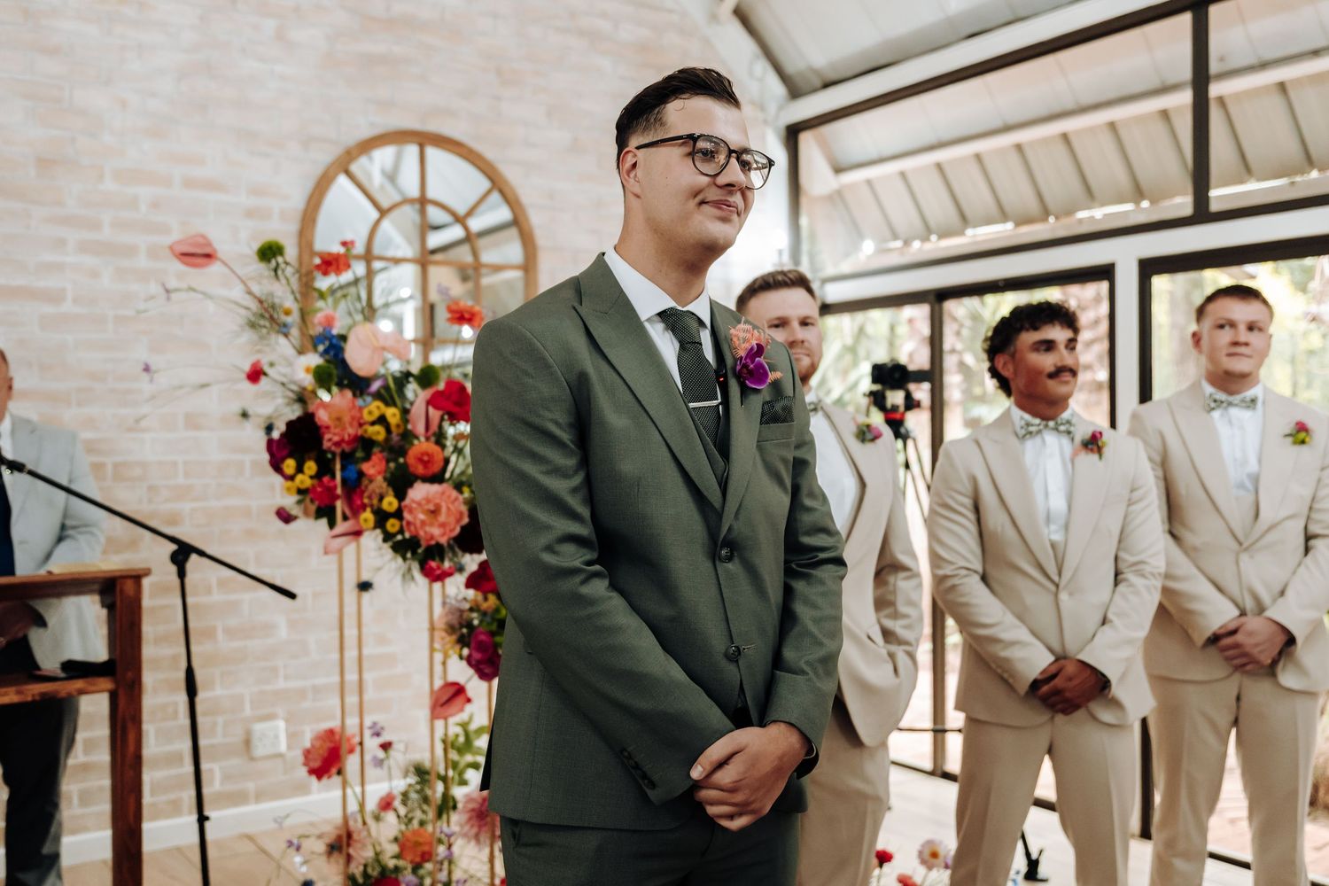Wedding party members in beige suits and olive green attire stand together during an elegant indoor ceremony.