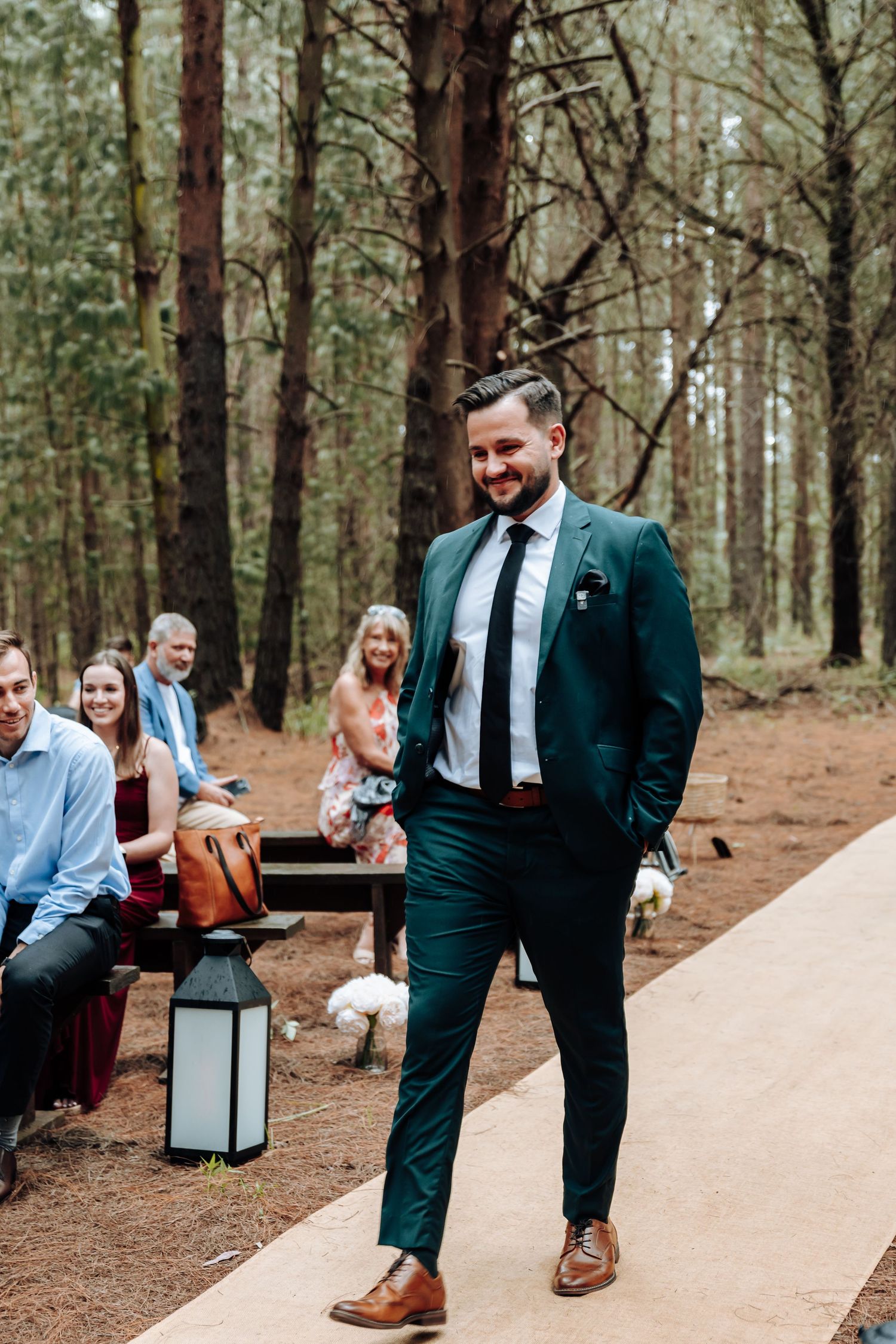 Wedding guests sit on wooden benches in a forest setting while groomsmen stand along a white aisle runner.