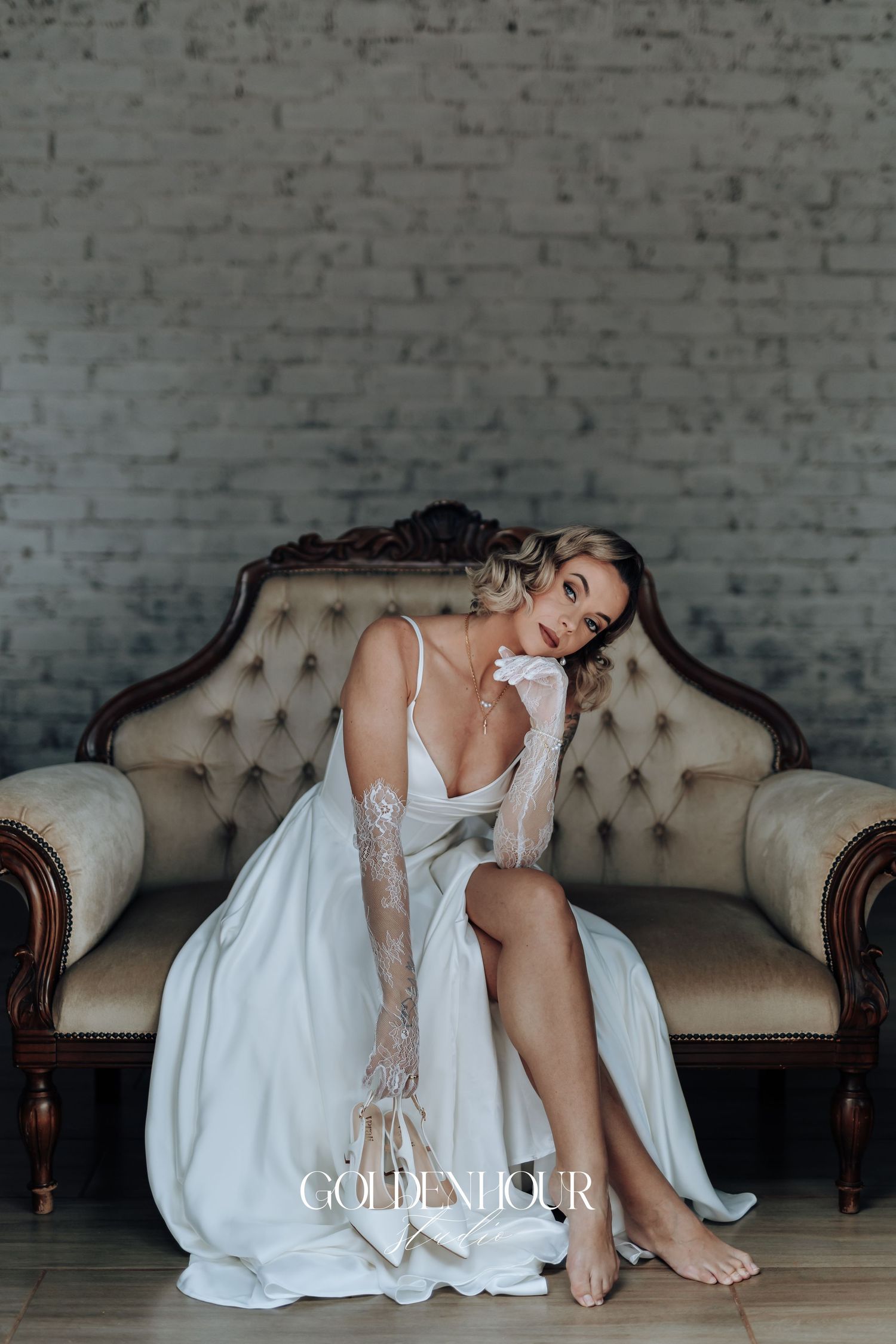 Bride in white dress reclining gracefully on a vintage tufted sofa against brick wall.