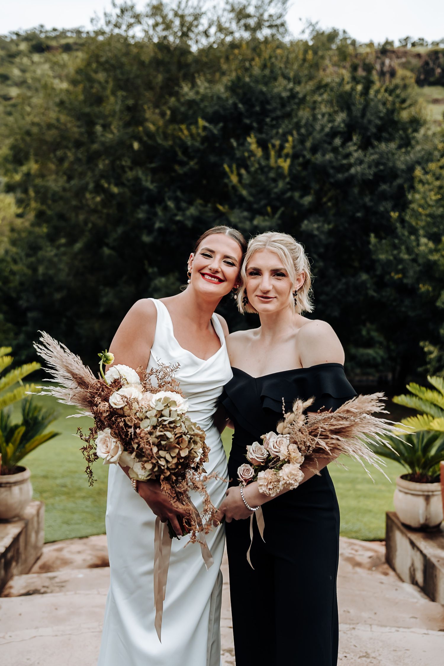 Two people in contrasting white and black formal attire share joyful moments while holding dried floral bouquets outdoors.