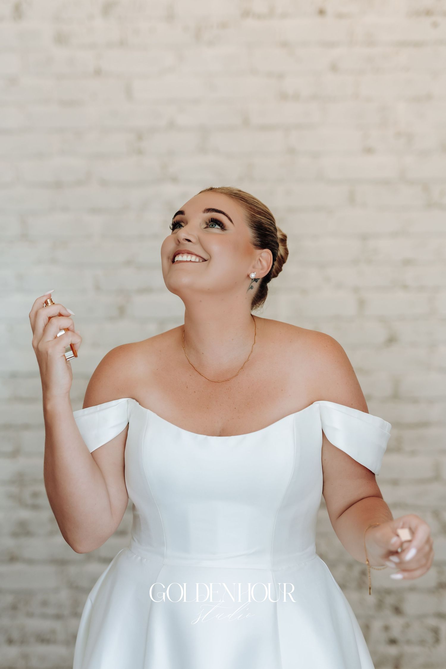 A joyful bride in an elegant off-shoulder white wedding gown smiles while looking up against a light brick wall.